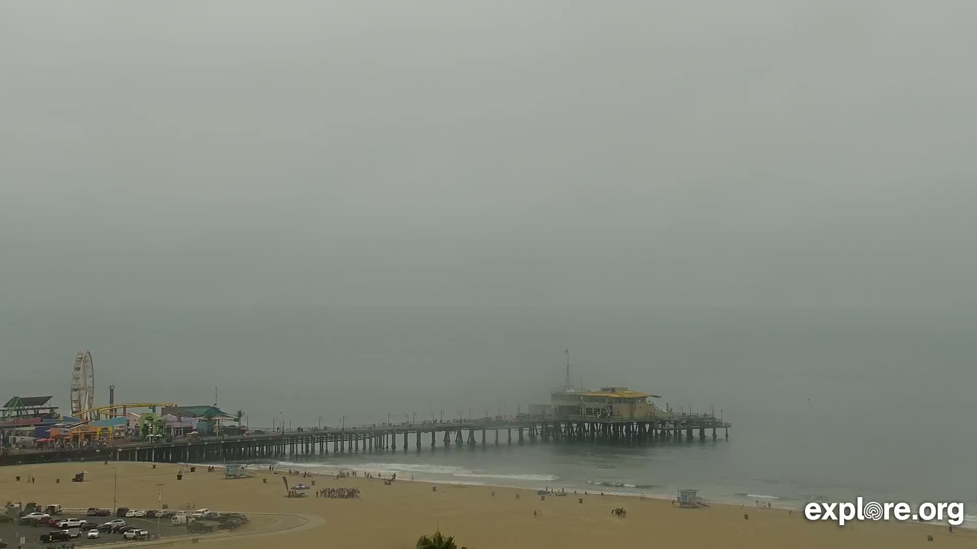 A hazy beach scene shows a pier extending from a sandy shore into a foggy ocean, with a Ferris wheel and other amusement park structures visible in the background.