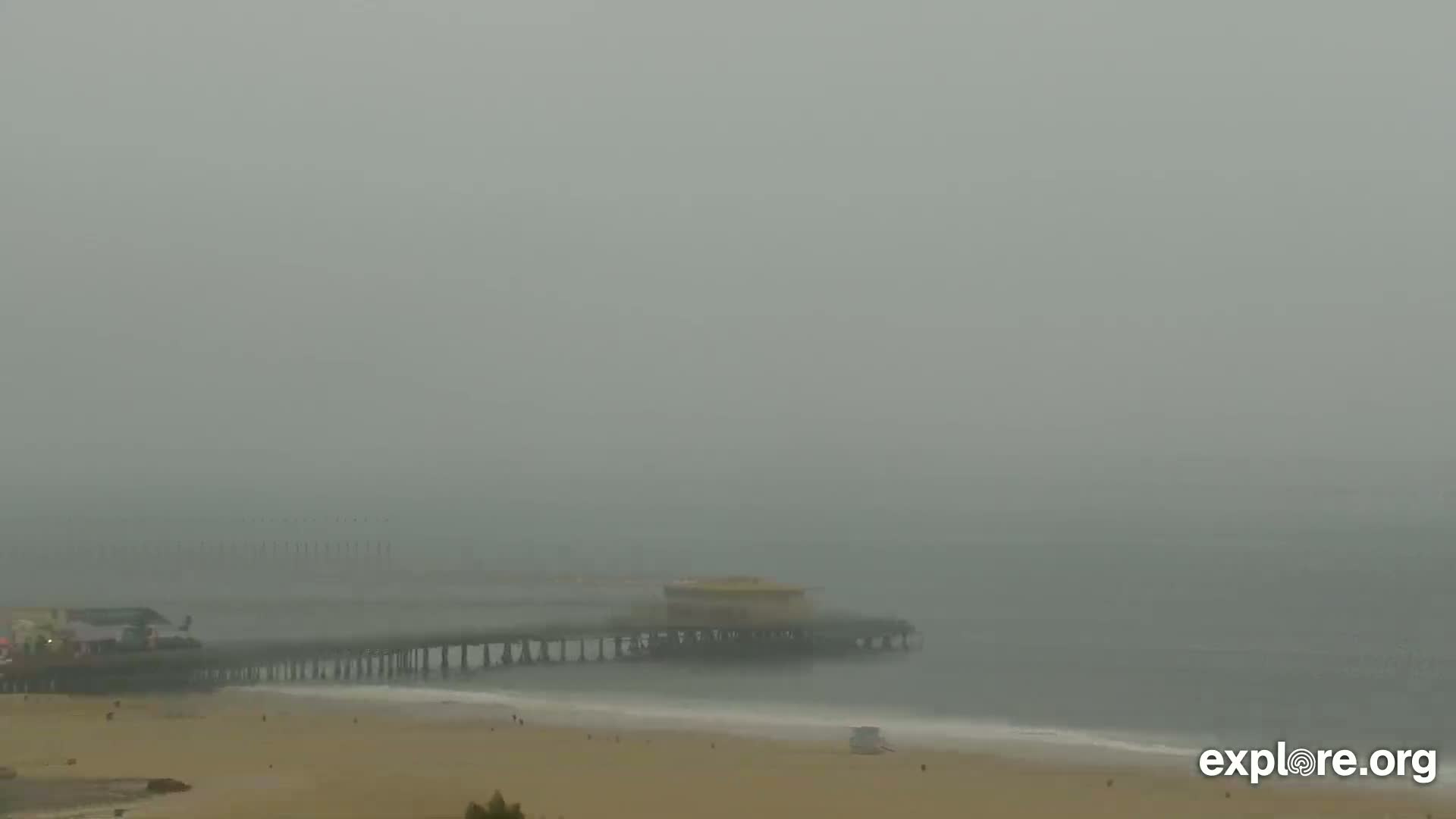 A long pier extends over a foggy ocean, with a sandy beach visible in the foreground.