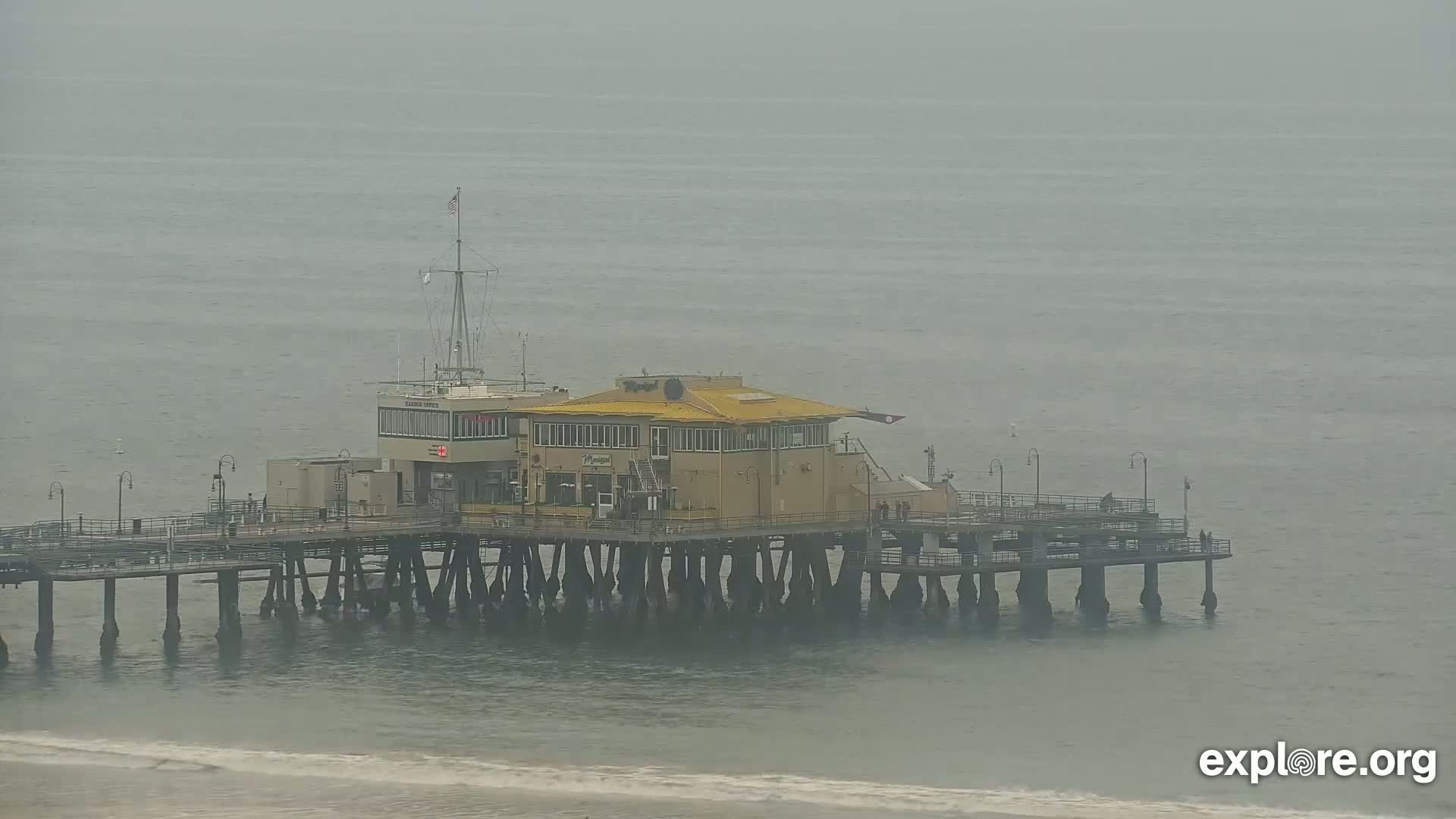 A long pier with a yellow building extends over a calm, gray ocean under an overcast sky.