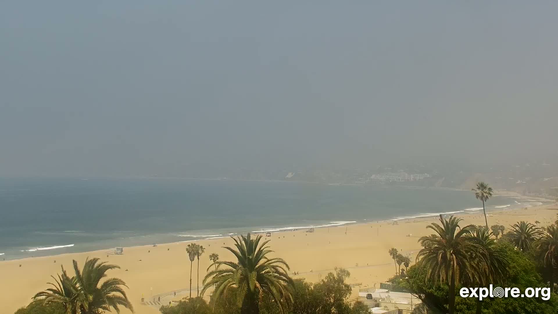 A hazy, sunny day shows a long sandy beach with palm trees and a calm ocean in the background.
