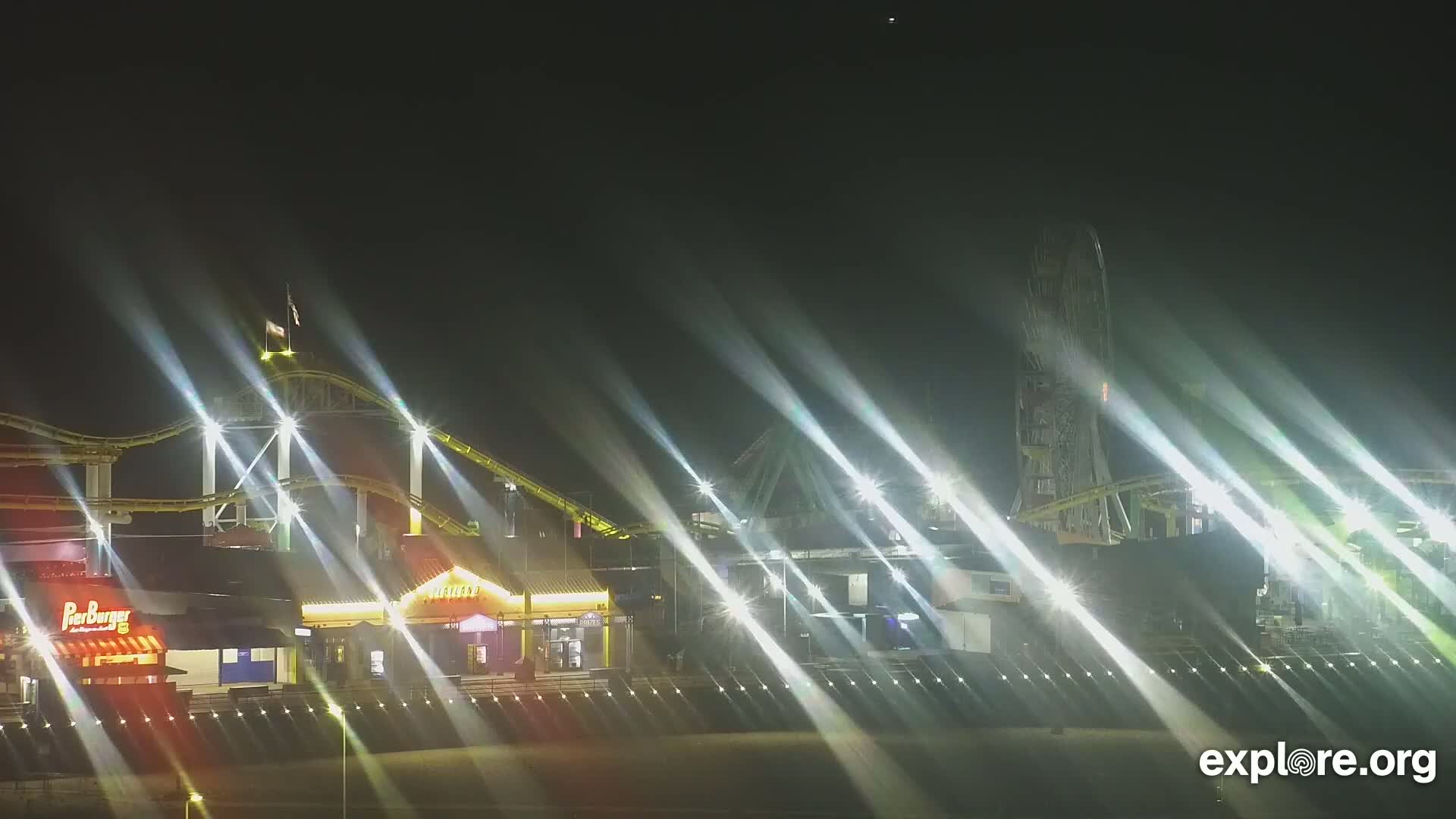 A nighttime view of an illuminated amusement park featuring a roller coaster and Ferris wheel, with bright light beams radiating across the scene.