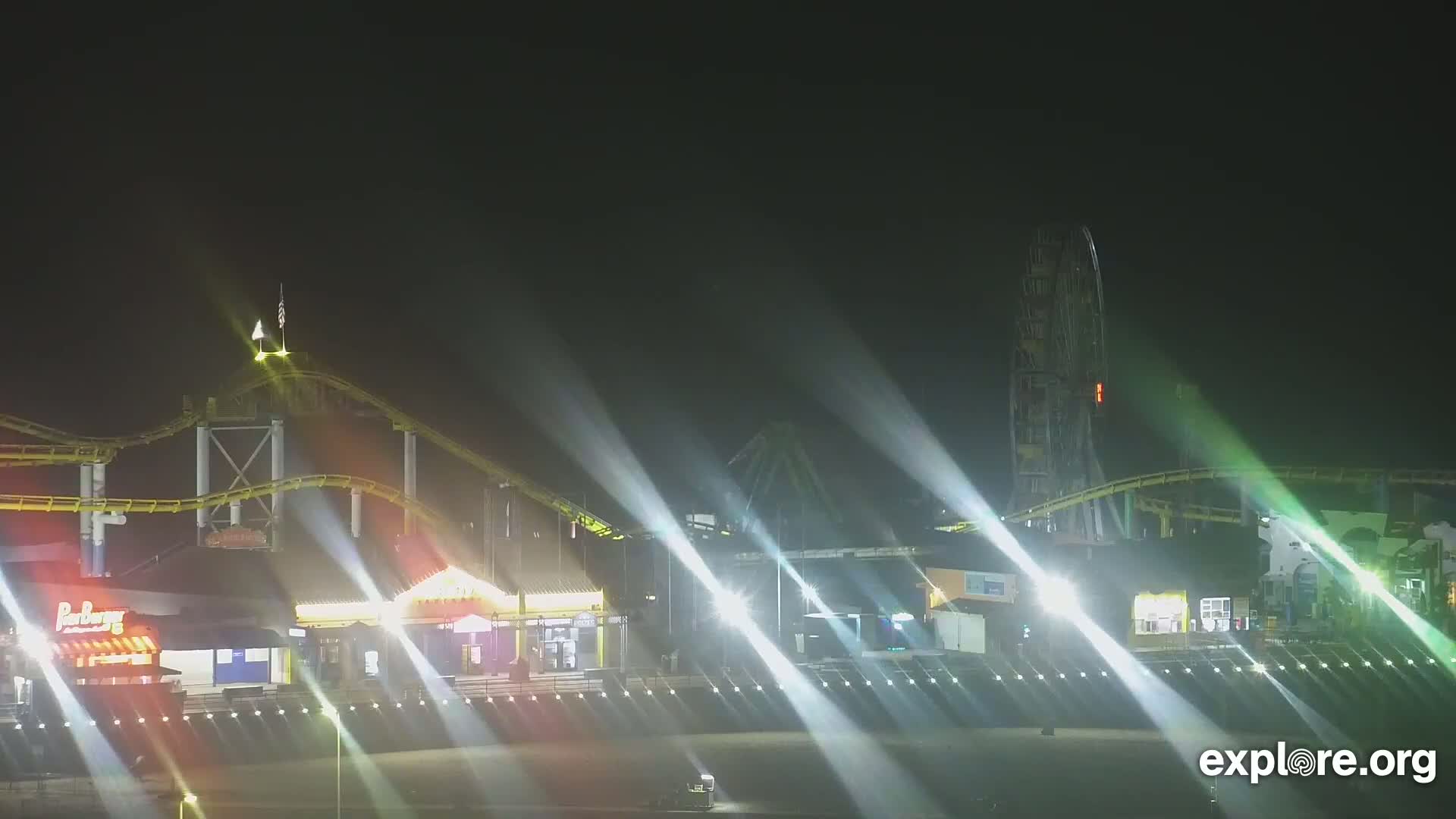 A brightly lit amusement park at night, featuring a roller coaster and Ferris wheel, is illuminated by strong spotlights under a dark sky.