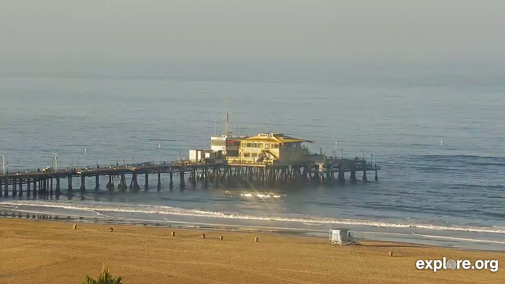 A long pier with a building on it extends into a calm ocean with a sandy beach in the foreground.
