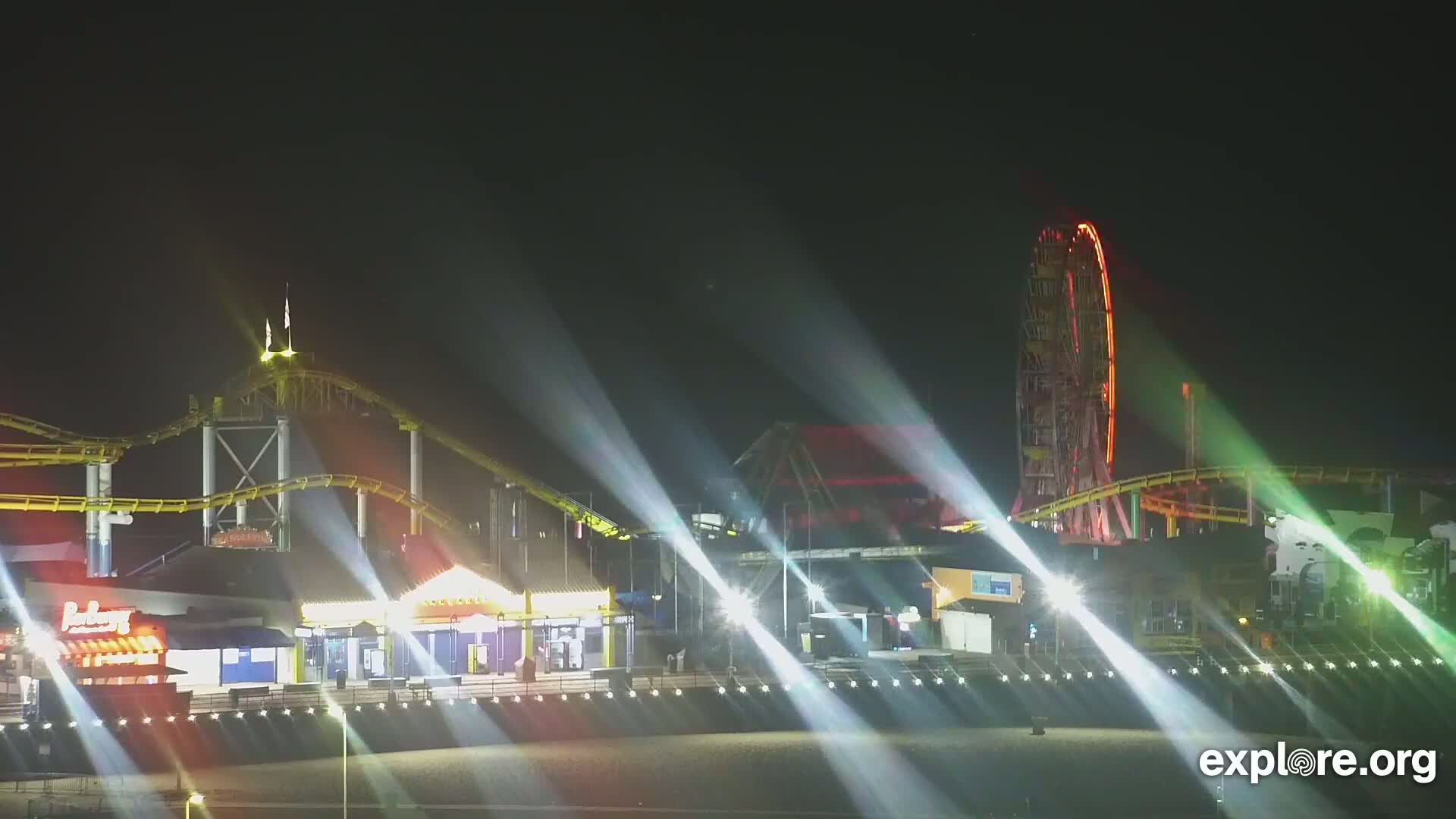 At night, brightly lit amusement park rides, including a roller coaster and Ferris wheel, are illuminated by searchlights on a beachfront boardwalk.