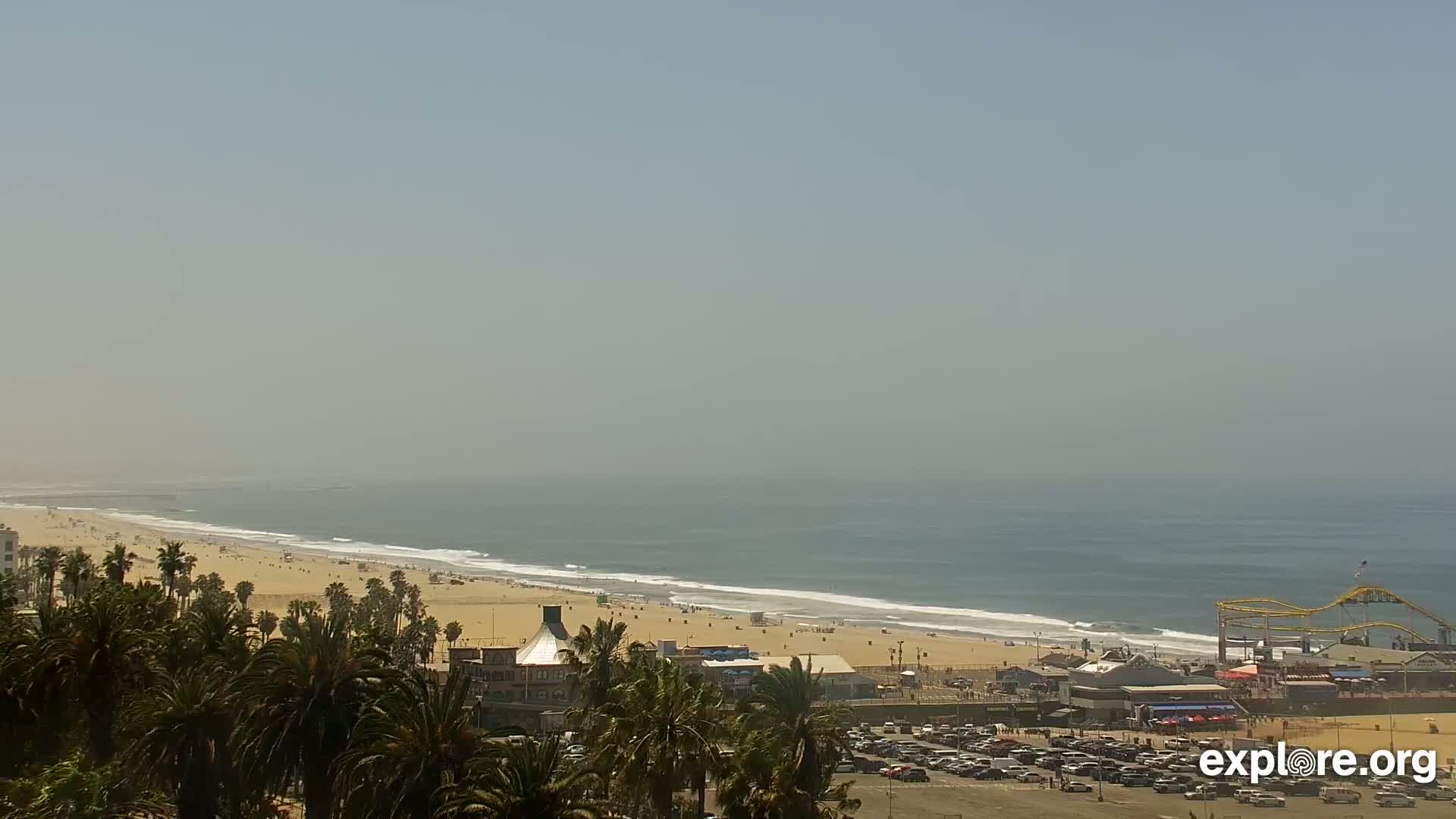 A hazy, sunny day reveals a long sandy beach with calm waves, palm trees, and a beachfront amusement park in the distance.