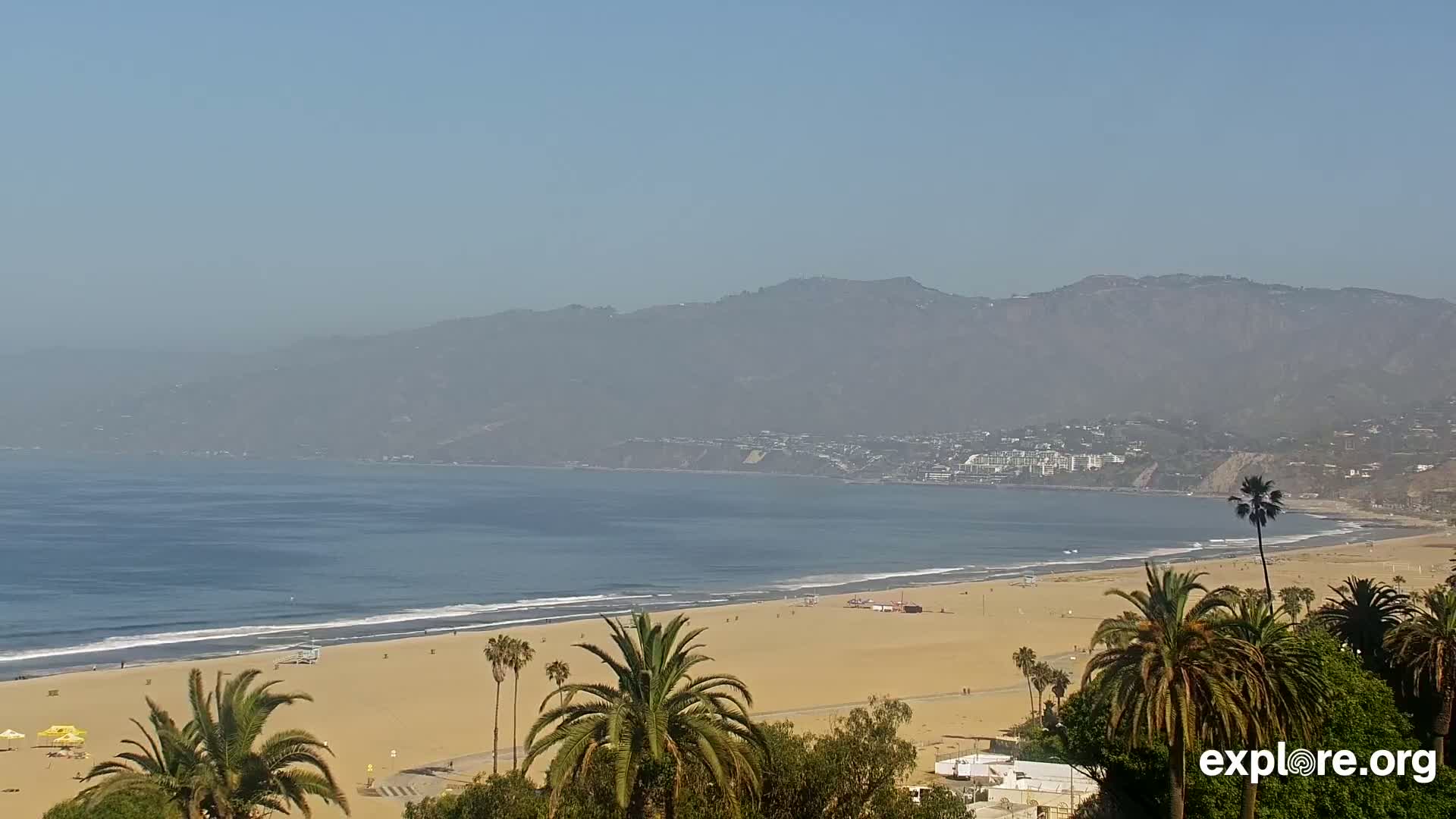 A hazy, sunny day overlooks a sandy beach with calm waves lapping the shore, palm trees in the foreground, and mountains in the distance.
