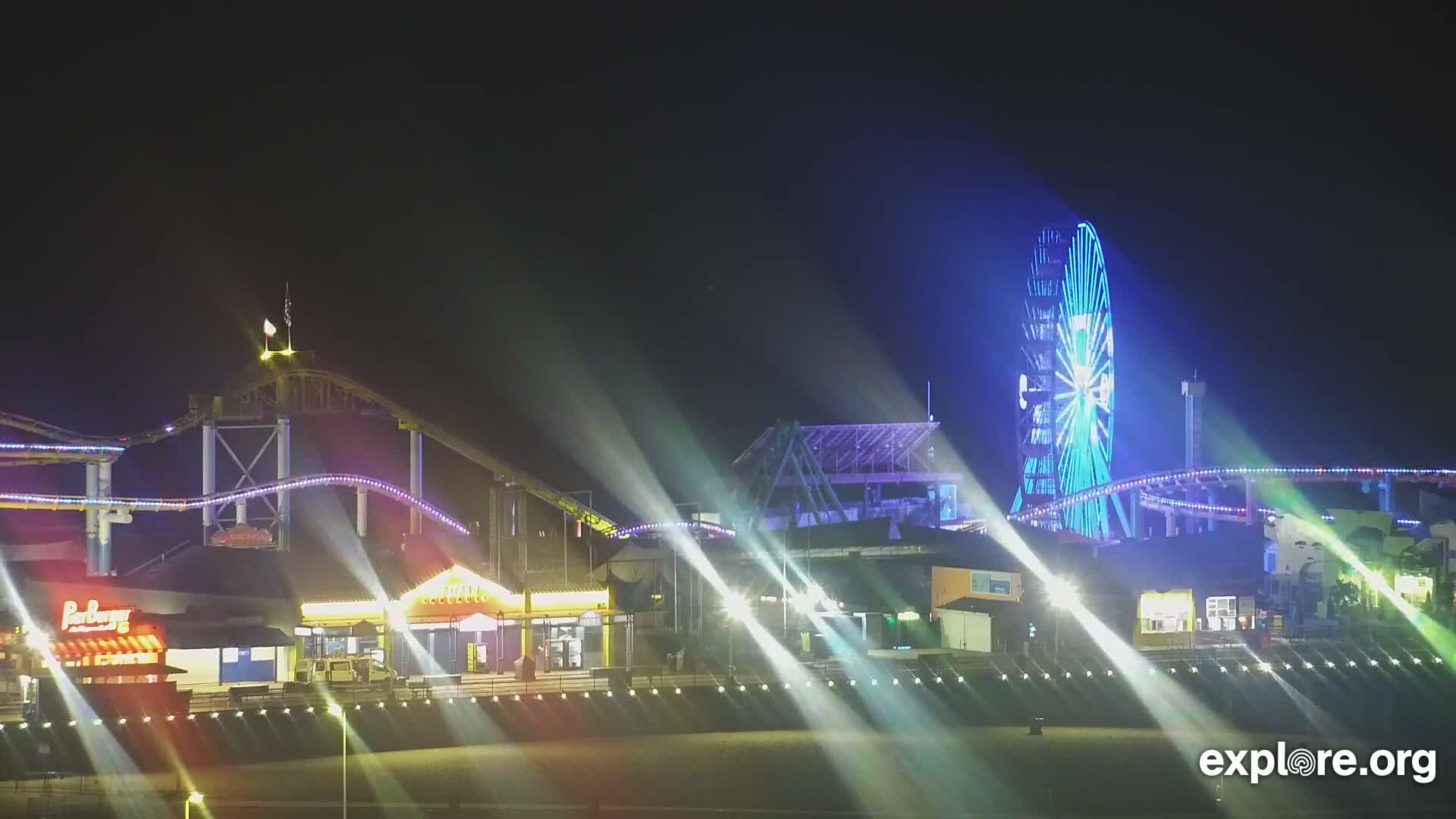 A brightly lit amusement park at night, featuring a Ferris wheel and roller coaster, is illuminated by spotlights.
