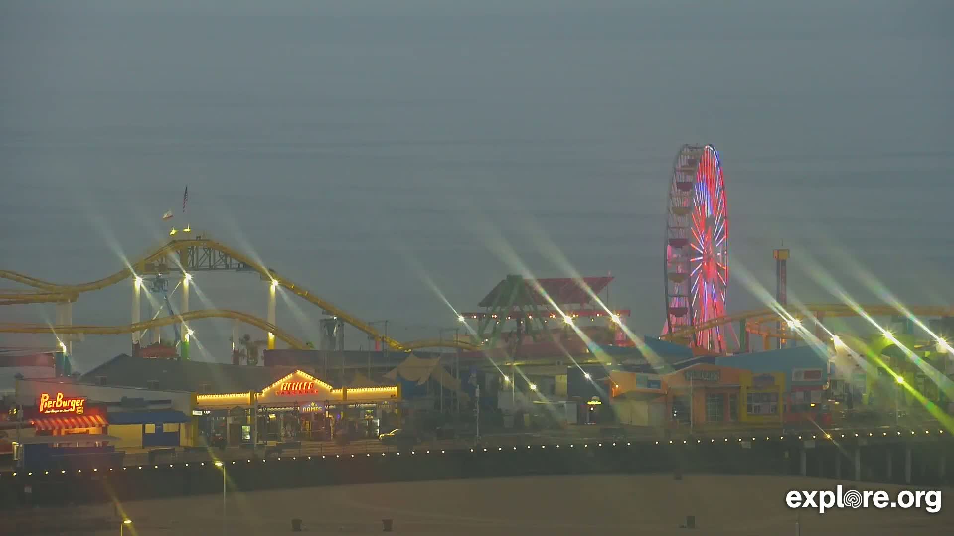 A brightly lit pier at night, featuring a roller coaster, Ferris wheel, and various shops, under a hazy sky.