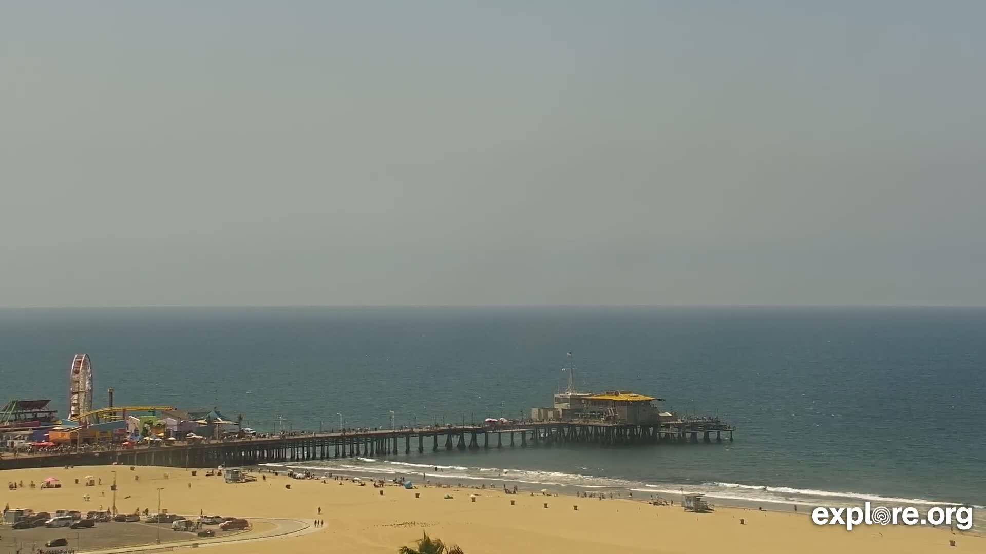 A long pier extends over a calm ocean with a sandy beach and amusement park structures visible in the background under a hazy sky.