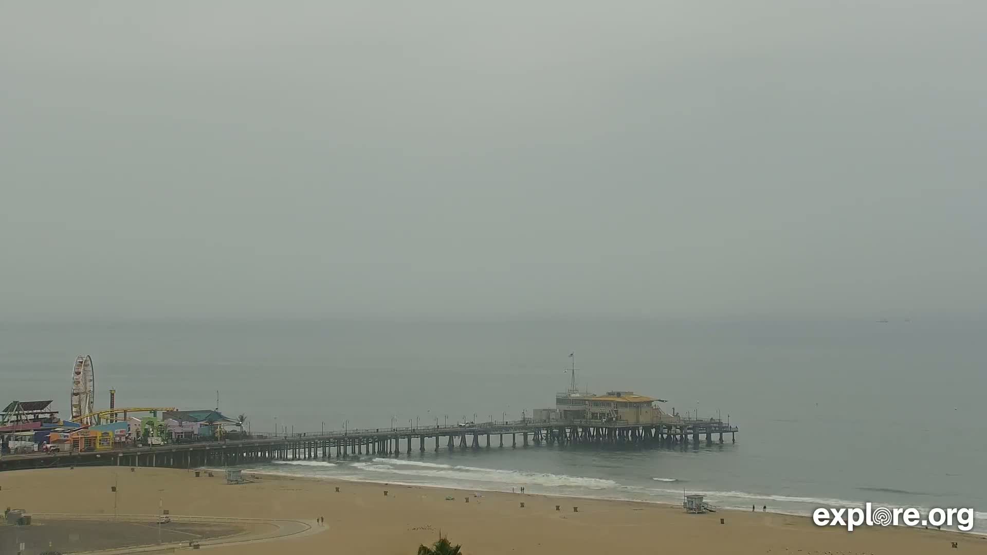A long pier extends from a sandy beach into a hazy, overcast ocean, with a Ferris wheel and other amusement park structures visible on the shore.