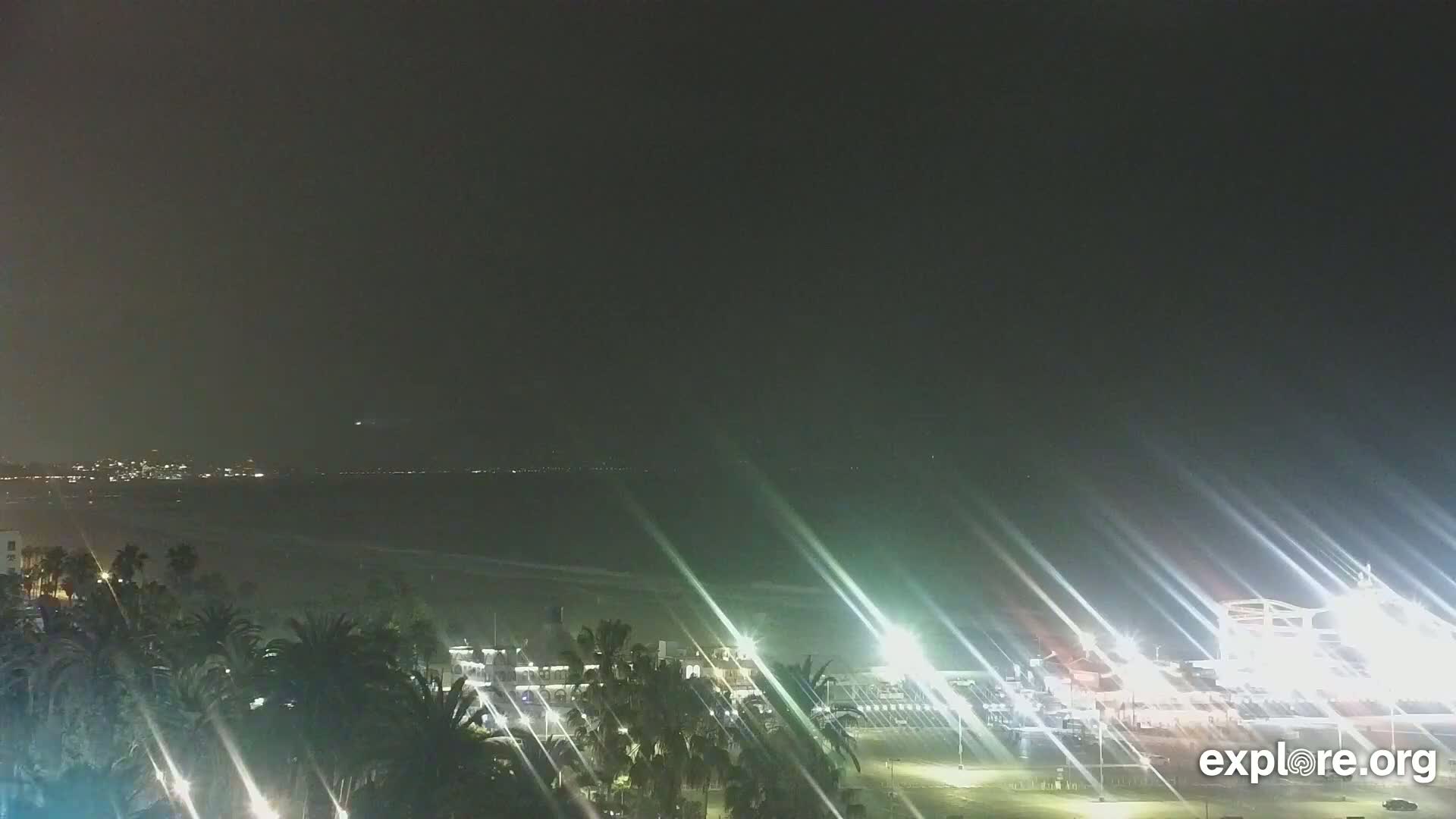 A nighttime coastal view shows a dark ocean, a city skyline in the distance, palm trees in the foreground, and brightly lit buildings and structures near the beach.