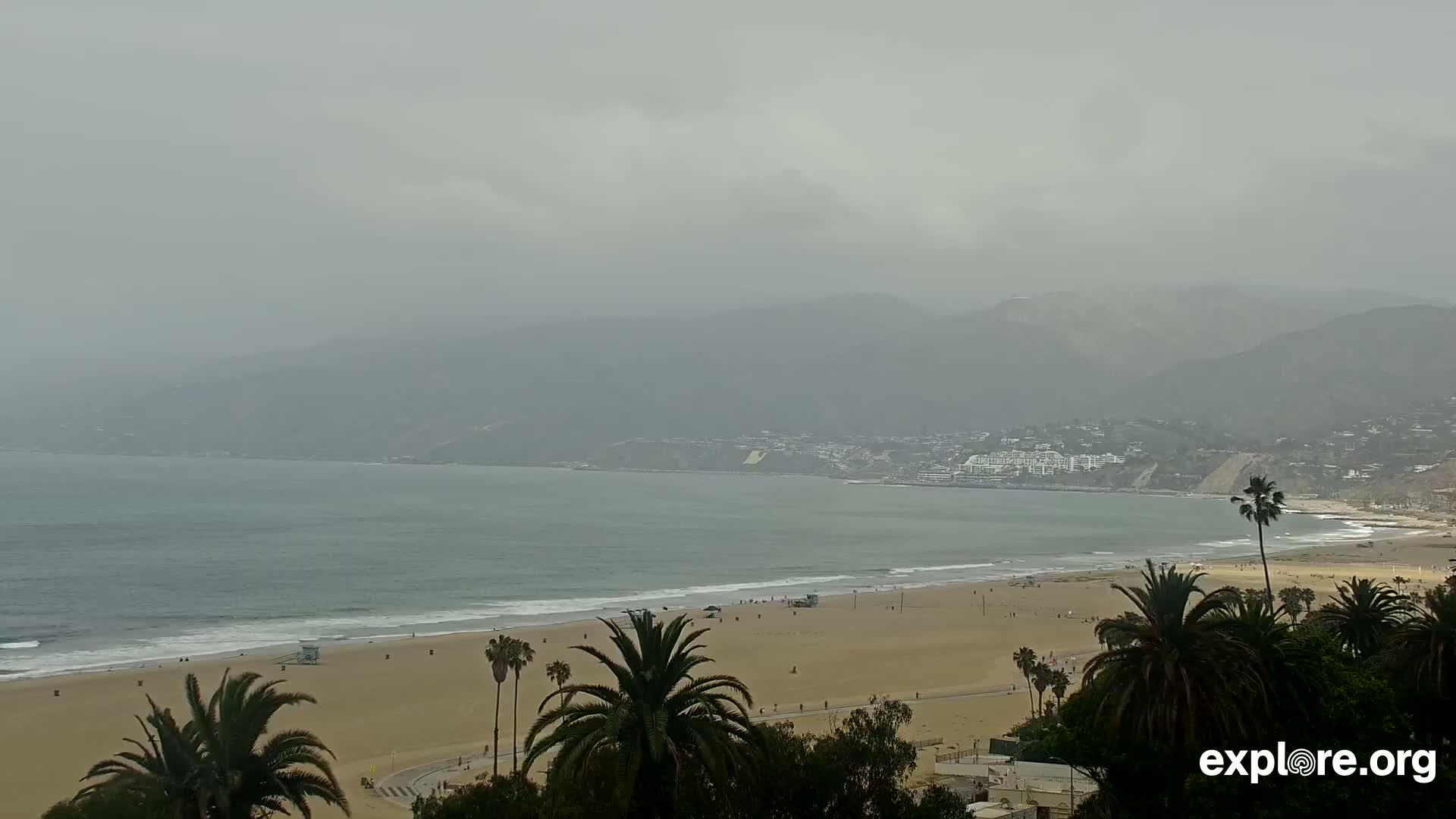 A hazy, overcast day reveals a sandy beach with palm trees, gentle waves lapping the shore, and distant mountains shrouded in mist.