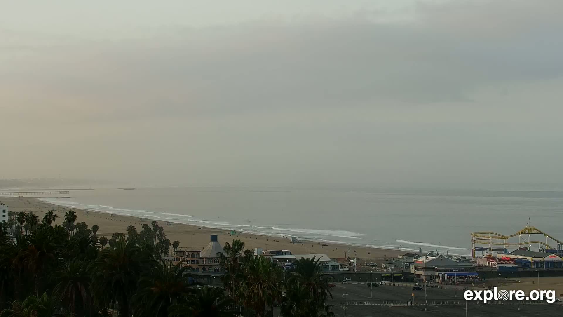 A hazy, overcast day reveals a long sandy beach with palm trees, buildings, and a pier with a roller coaster in the distance, all overlooking a calm ocean.