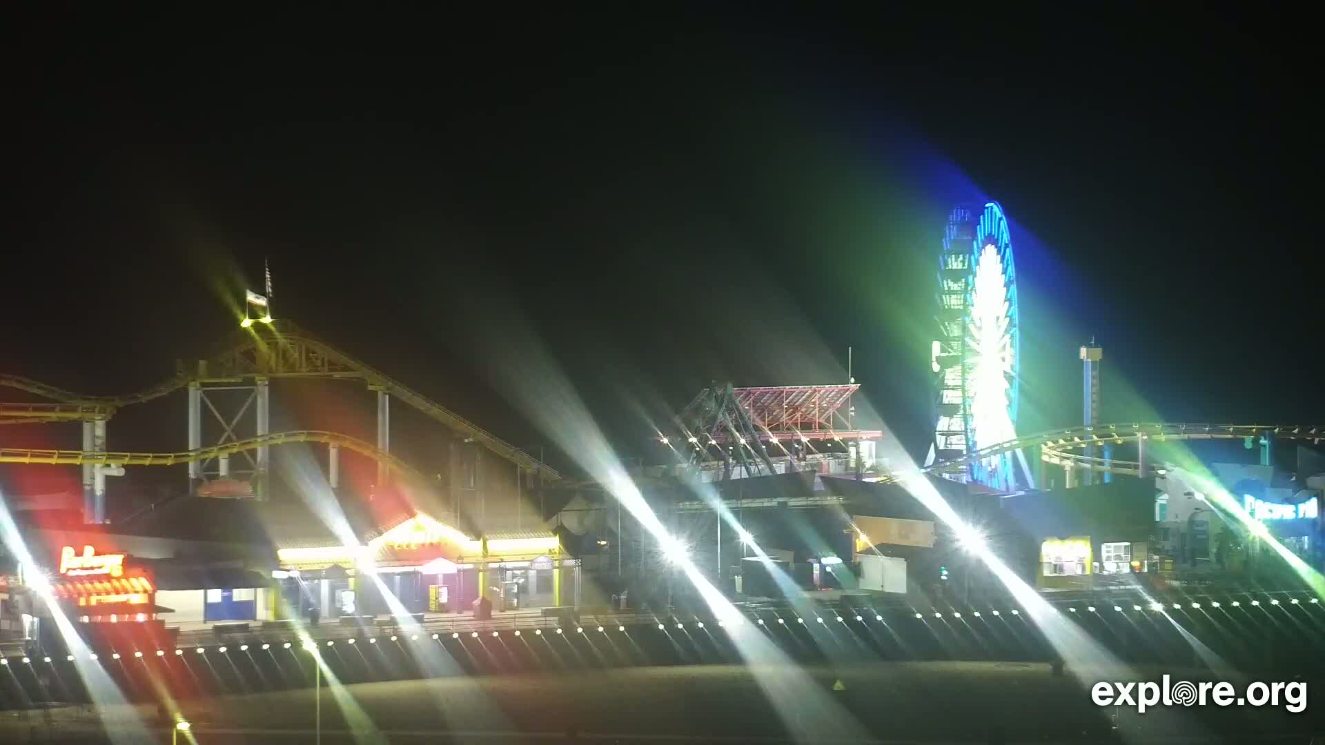 A brightly lit amusement park at night, featuring roller coasters and a Ferris wheel, is illuminated by searchlights.