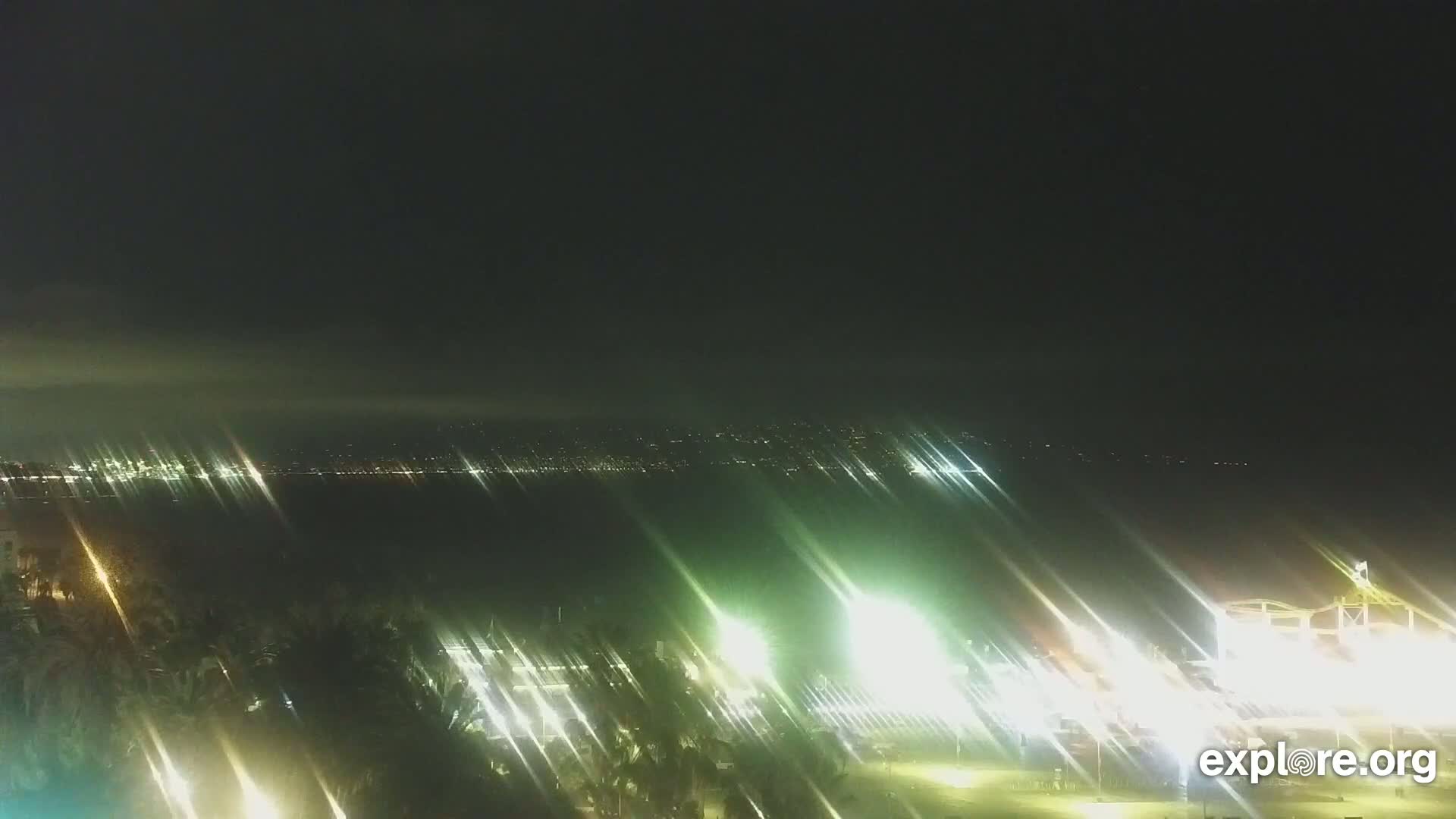 A nighttime view shows a brightly lit city across a body of water, with palm trees and lights in the foreground under a dark, possibly hazy sky.