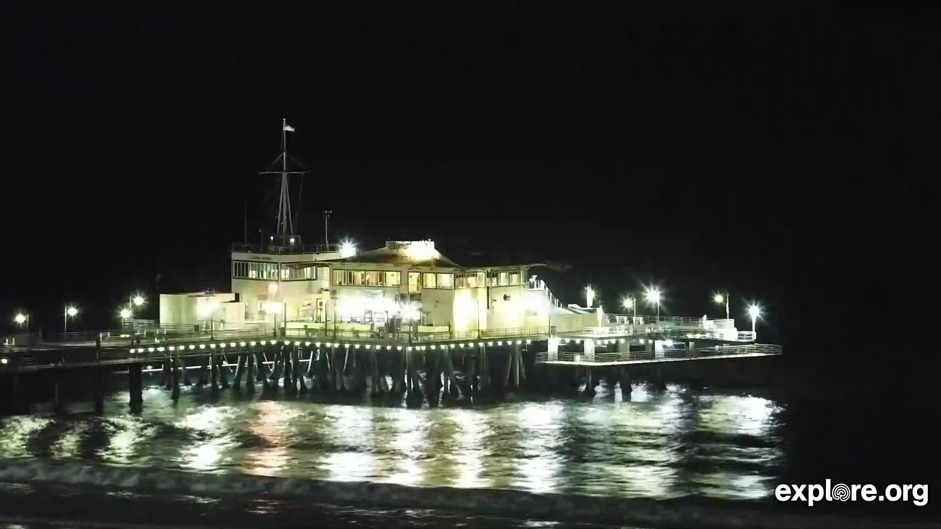 A well-lit pier building extends over the ocean at night, with bright reflections shimmering on the water under a clear, dark sky.
