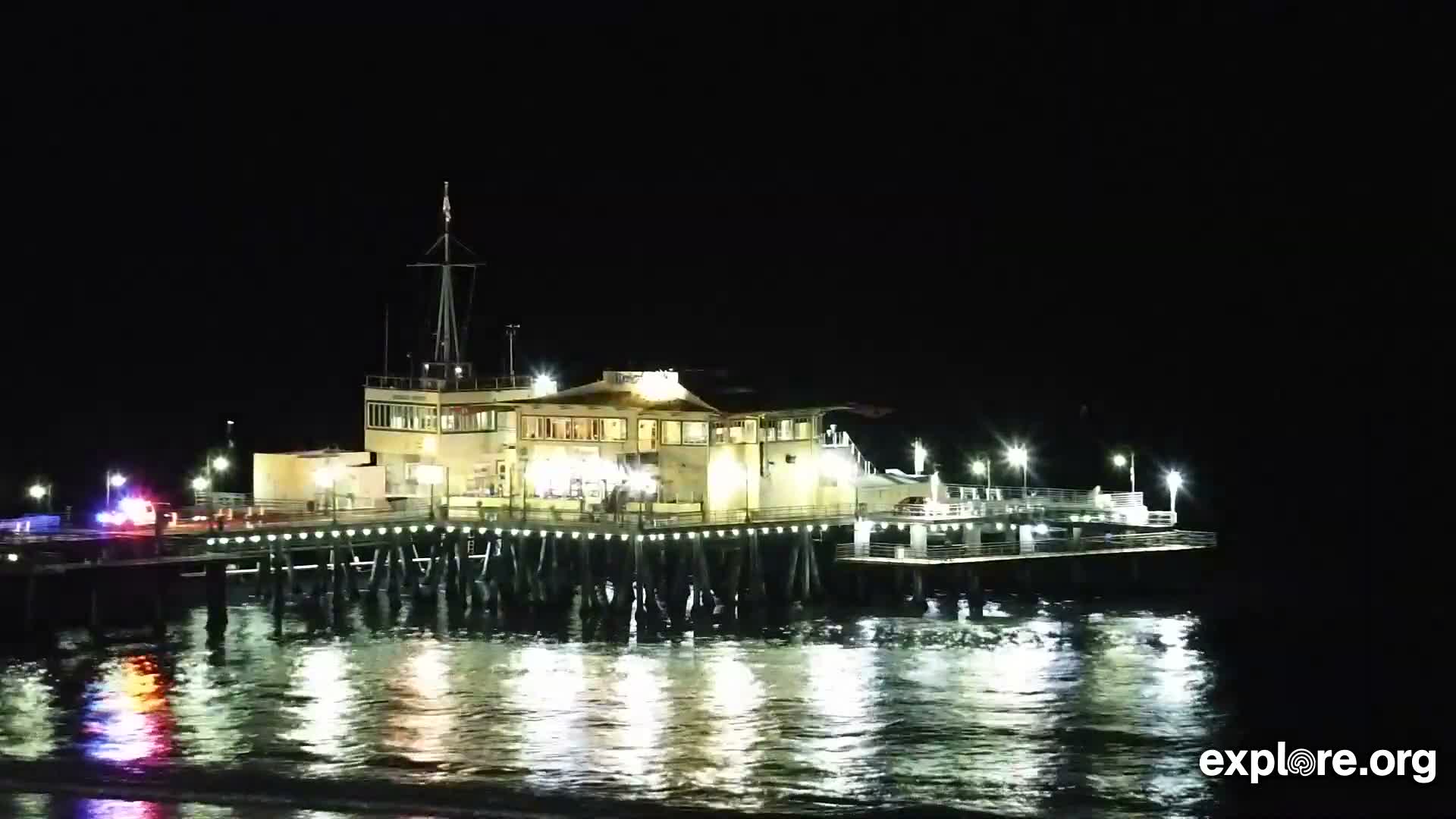 An illuminated pier featuring multiple buildings and a vehicle with flashing red and blue lights extends over calm, dark water reflecting the bright lights, all under a clear night sky.