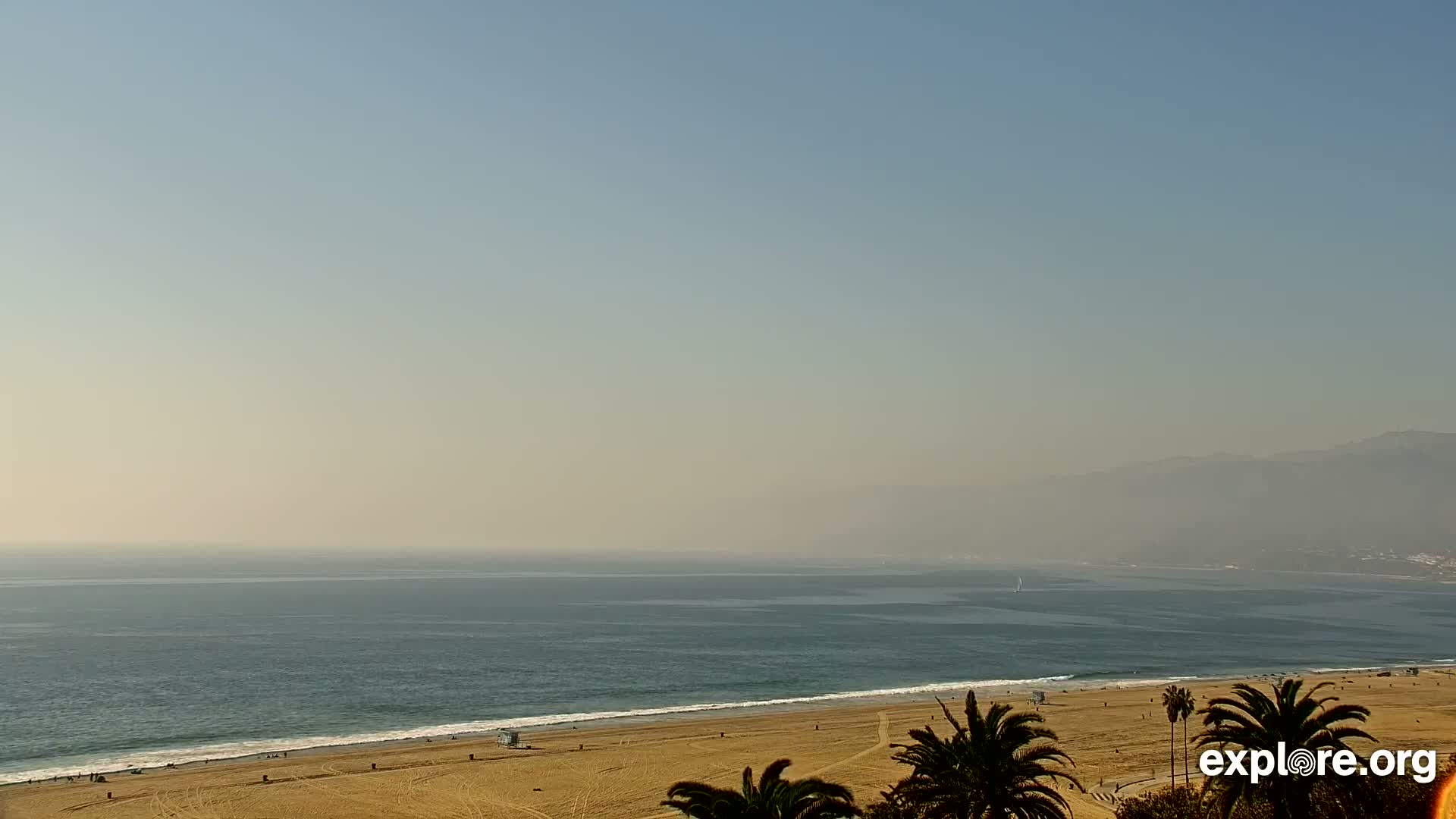 A wide sandy beach with palm trees and a lifeguard stand borders a calm blue ocean with a visible sailboat, all under a hazy, bright sky revealing distant mountains along the coast.