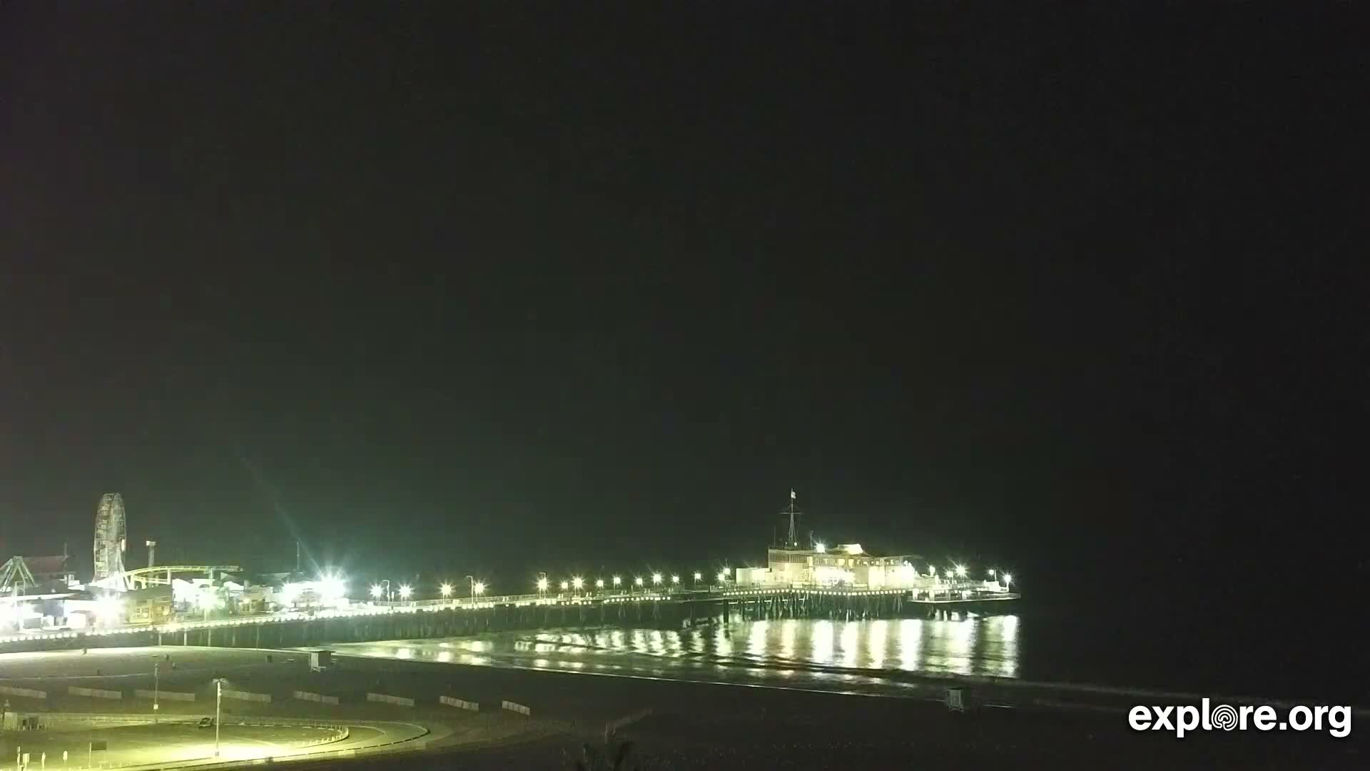 A brightly lit amusement pier, featuring a Ferris wheel and various buildings, stretches over a dark ocean and sandy beach on a clear night.