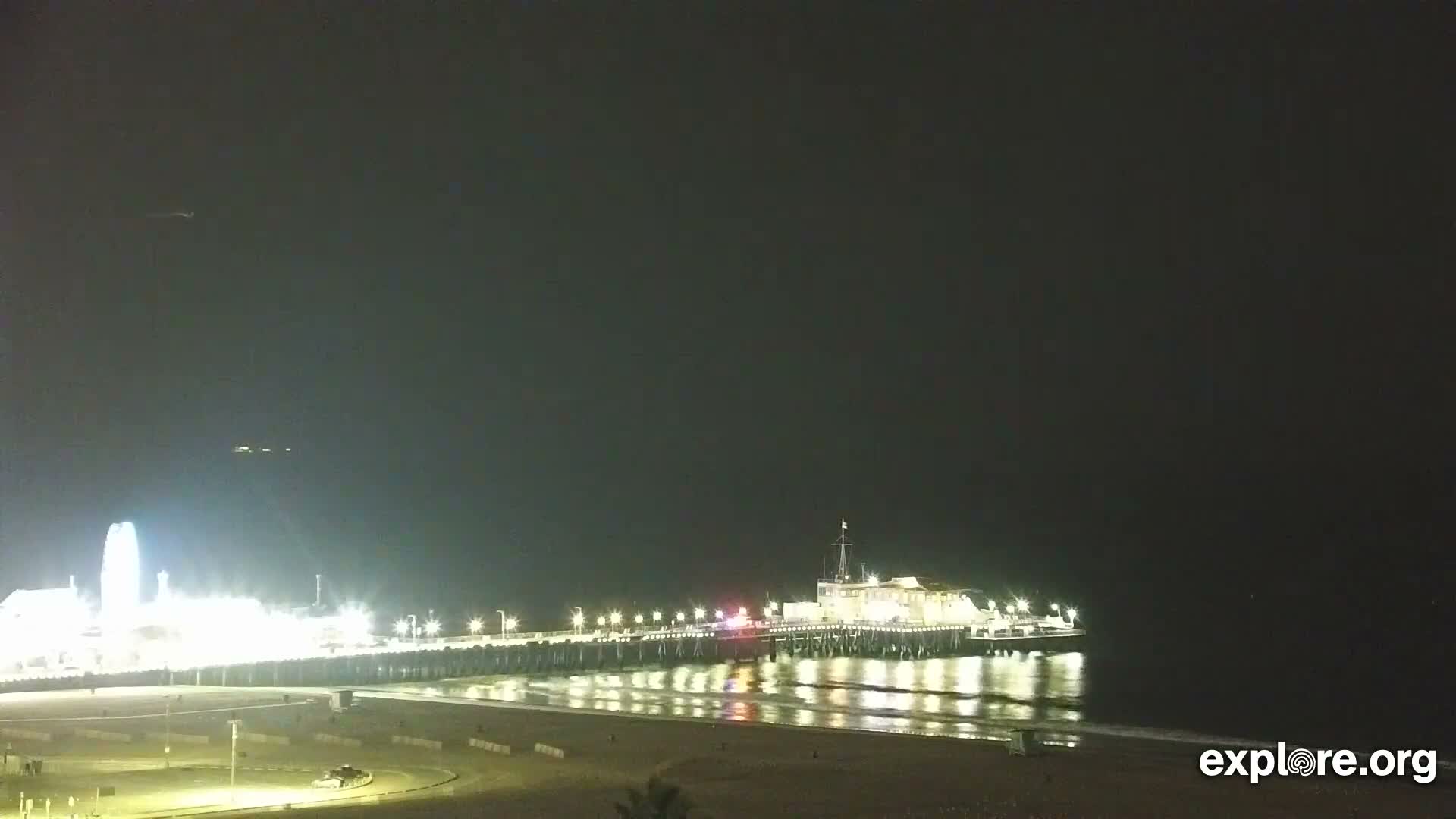 A brightly lit pier featuring amusement park rides and structures extends over calm dark water next to a sandy beach under a clear night sky.