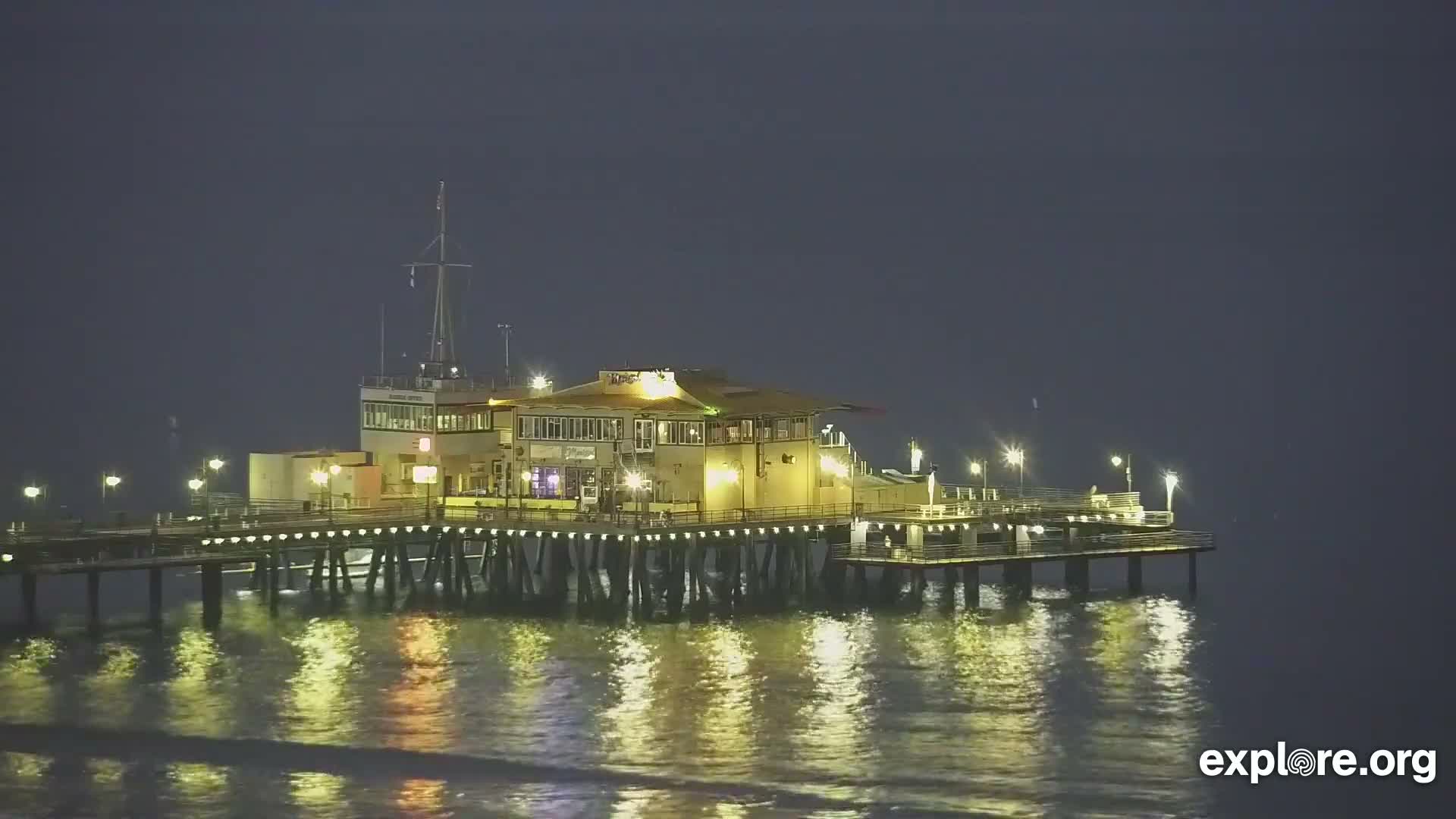 A brightly lit pier featuring amusement park rides and structures extends over calm dark water next to a sandy beach under a clear night sky.