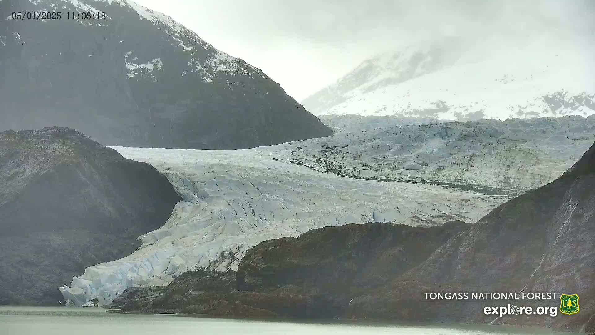 A large glacier flows down between dark, rocky mountains on an overcast day.