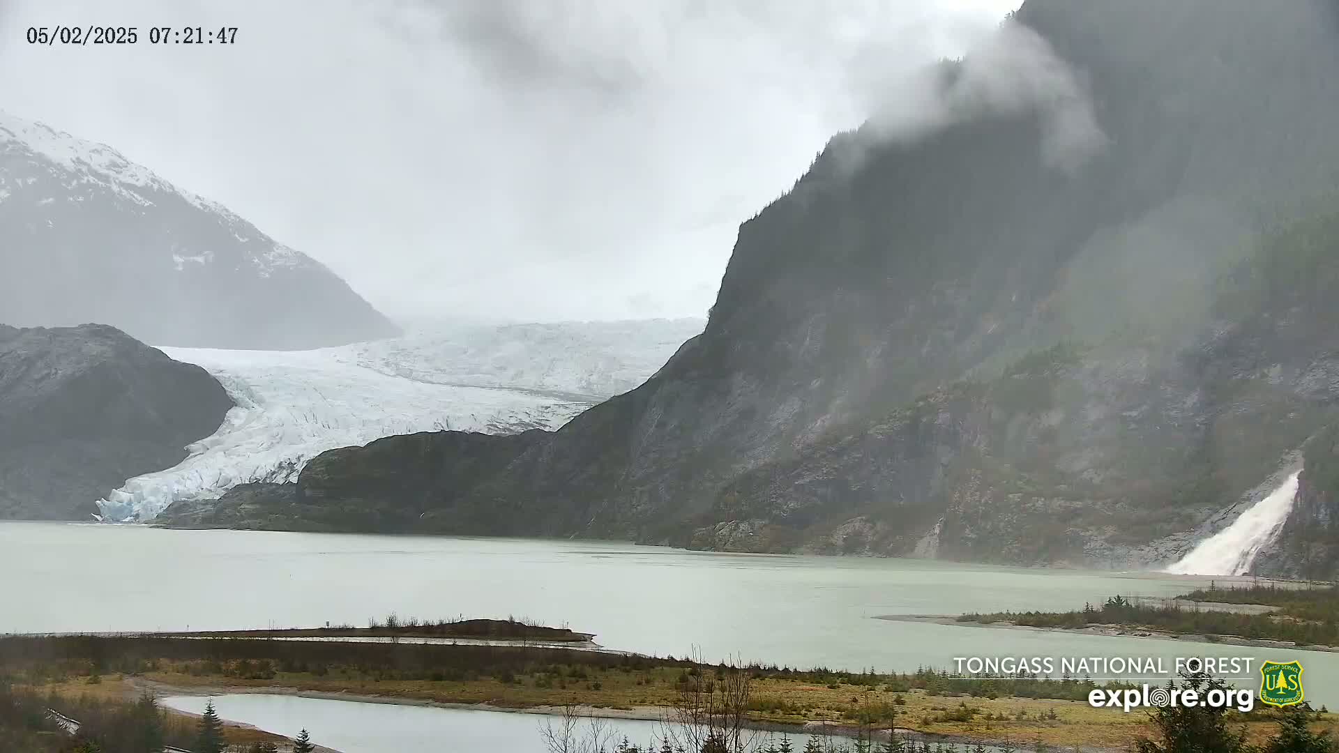 A glacier flows into a milky-green lake surrounded by misty, dark mountains under an overcast sky.