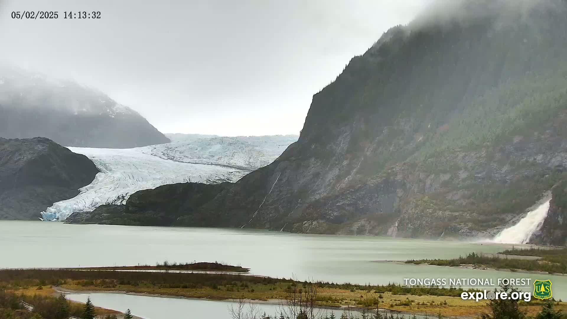 A glacier flows into a milky-green lake under a cloudy, rainy sky, with waterfalls cascading down a dark mountainside.