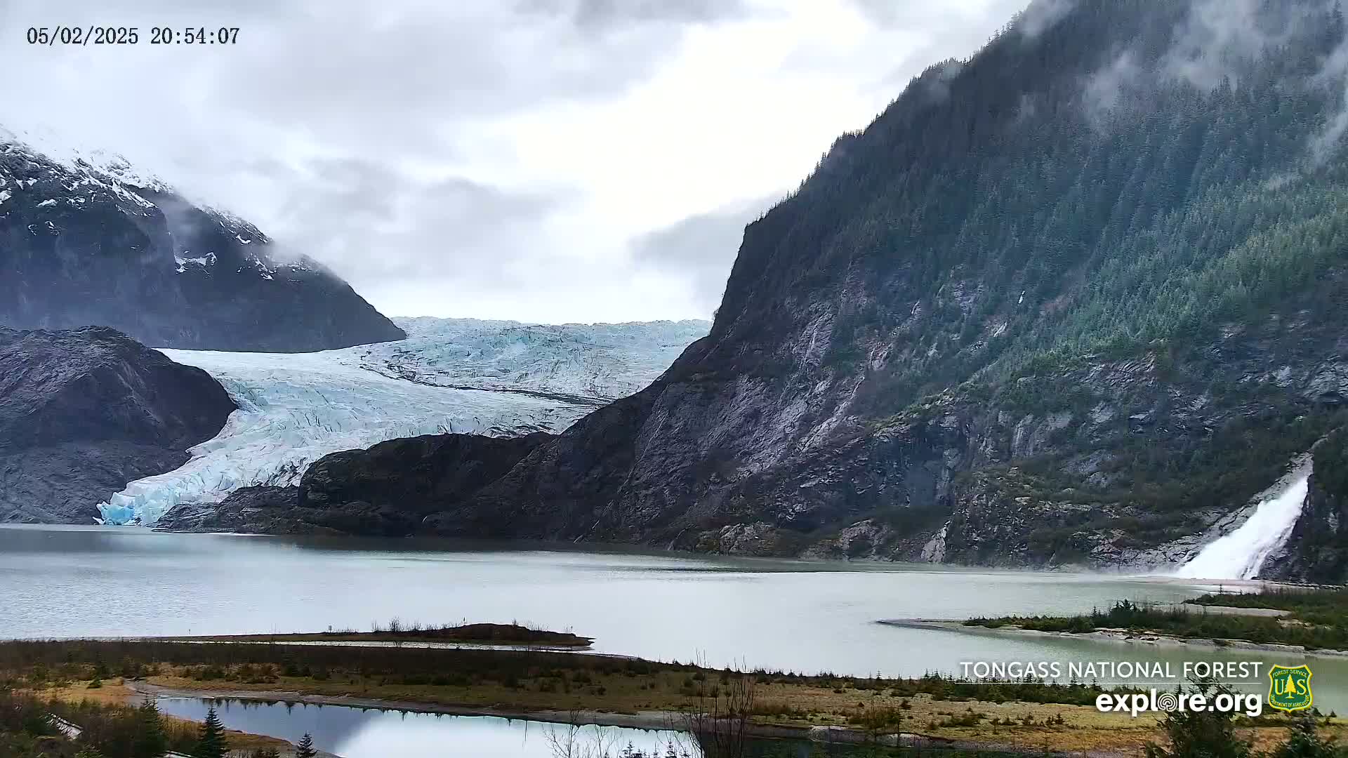 A glacier flows into a calm lake nestled between steep, dark mountains under an overcast sky.