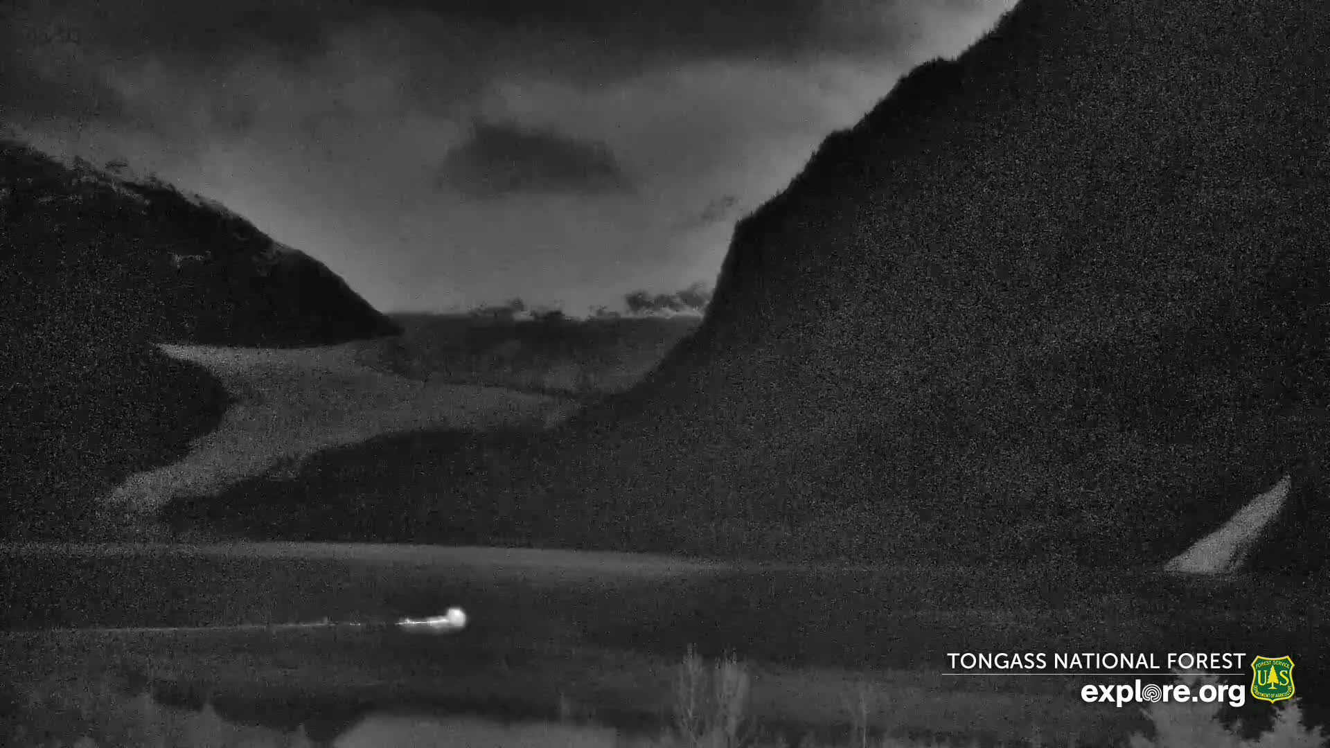 A dark, overcast scene shows a valley between two large, dark mountains, with a faint light visible in the valley below.