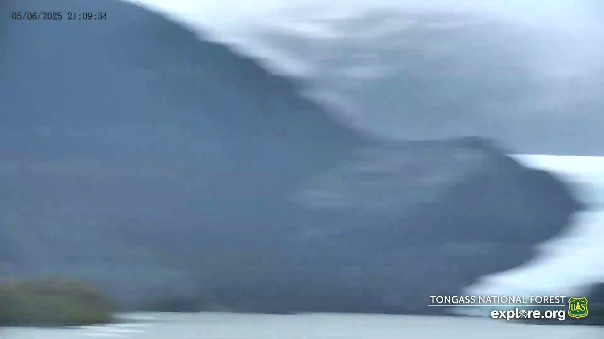 A blurry image shows a dark-colored mountainside next to a body of water under an overcast sky.
