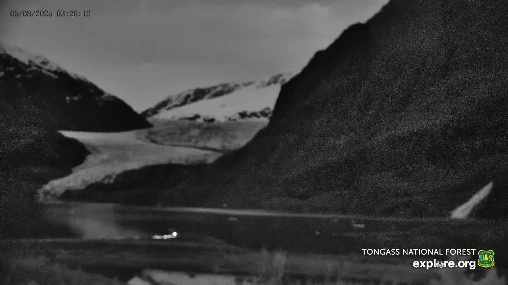 A glacier is visible between dark, mountainous terrain under a cloudy, nighttime sky, with a road and faint headlights in the foreground.