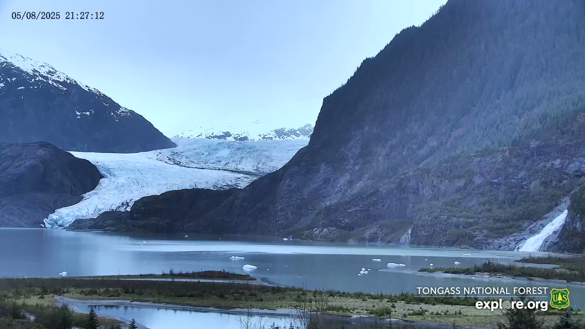 A large glacier flows into a calm lake nestled between dark, steep mountains under an overcast sky.