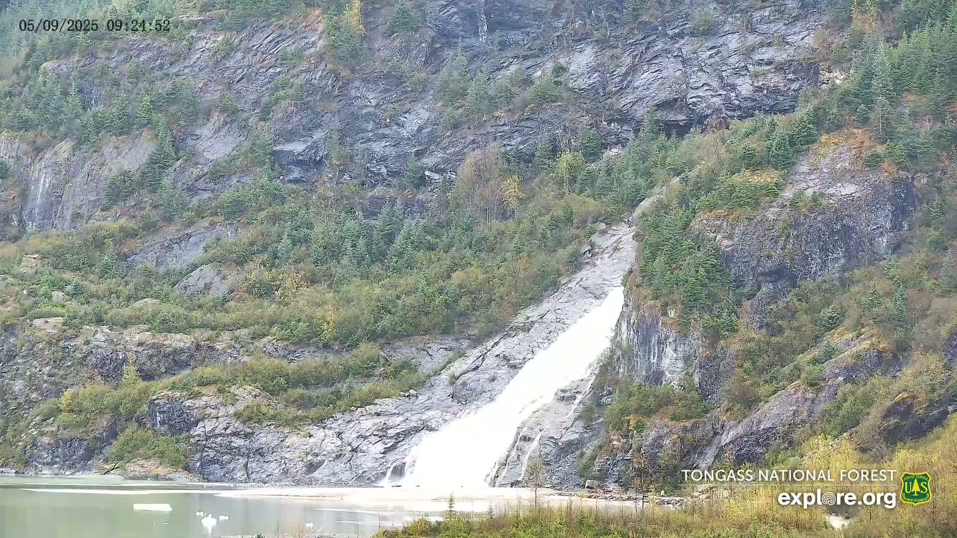 A waterfall cascades down a rocky, forested hillside into a calm body of water under an overcast sky.