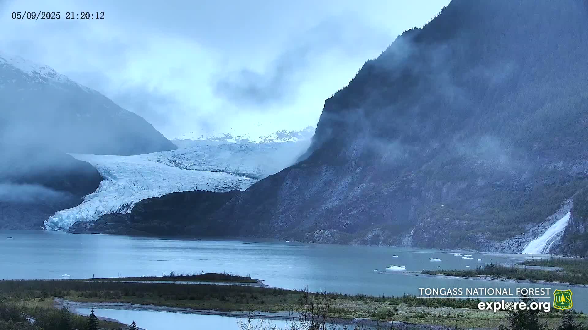 A glacier flows into a calm lake surrounded by misty mountains, one of which has a waterfall cascading down its side.