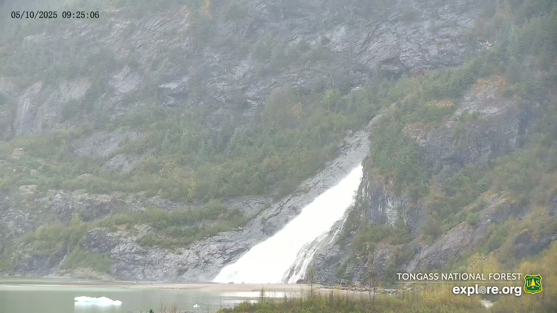 A large waterfall cascades down a rocky, green hillside into a body of water under an overcast sky.