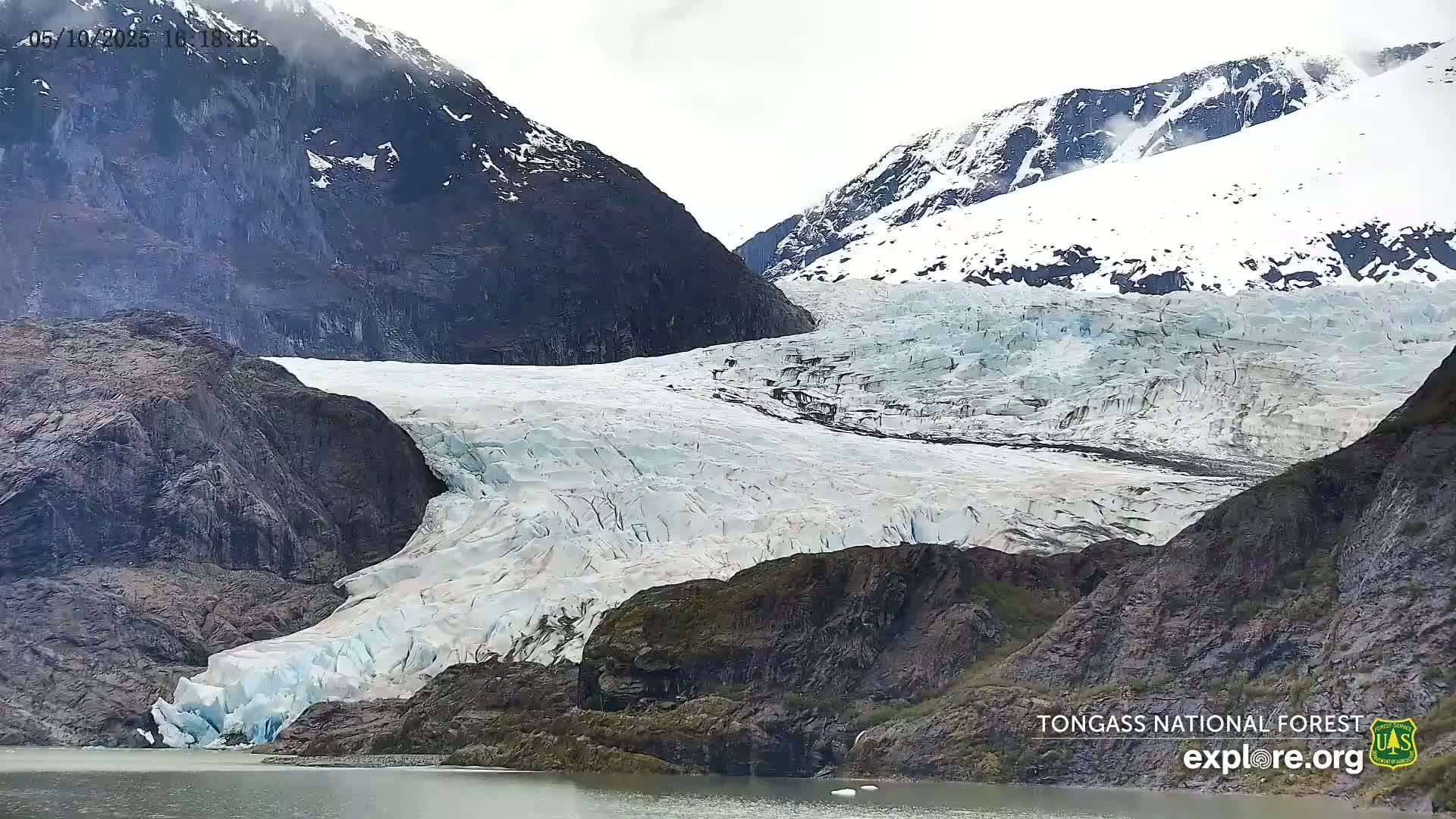 A large glacier flows down between dark, rocky mountains on an overcast day.