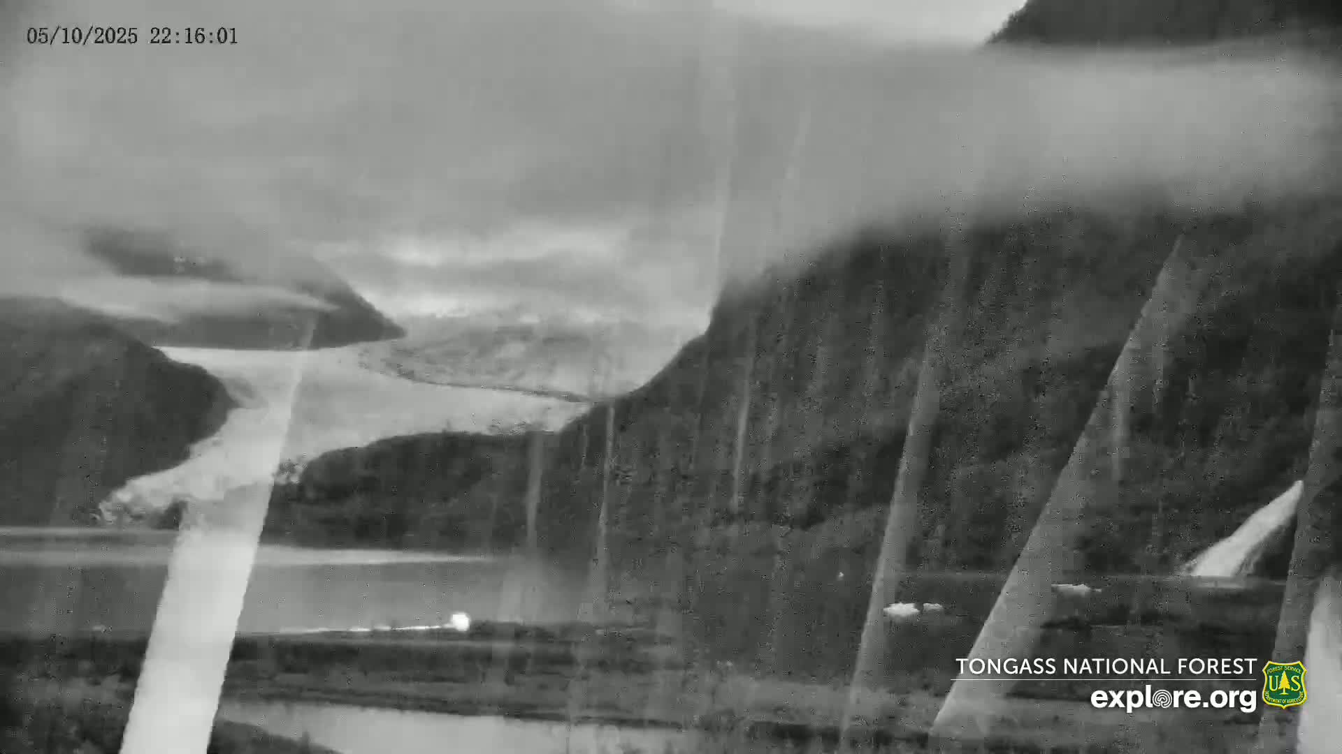 A grayscale image shows a glacier and body of water, partially obscured by fog and rain, with steep, dark mountains in the background.