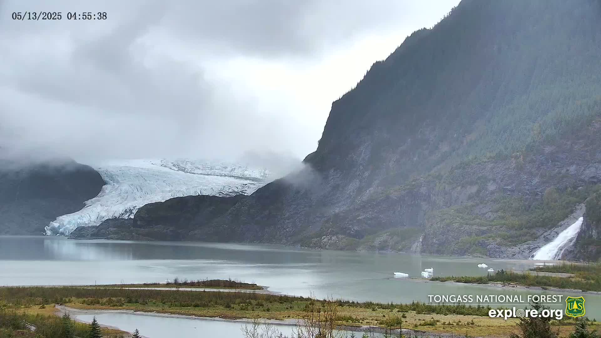 A glacier is partially obscured by fog next to a steep, dark mountain with a waterfall cascading into a calm lake on an overcast day.