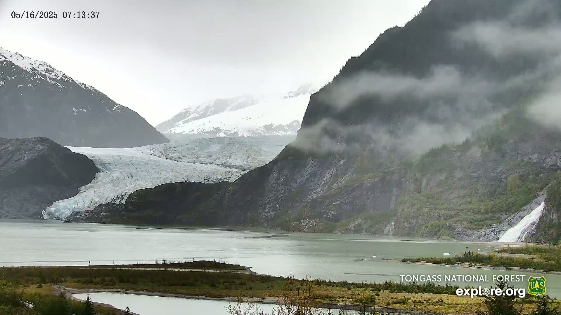 A glacier flows into a calm, cloudy body of water nestled between misty, snow-capped mountains, one of which features a waterfall.