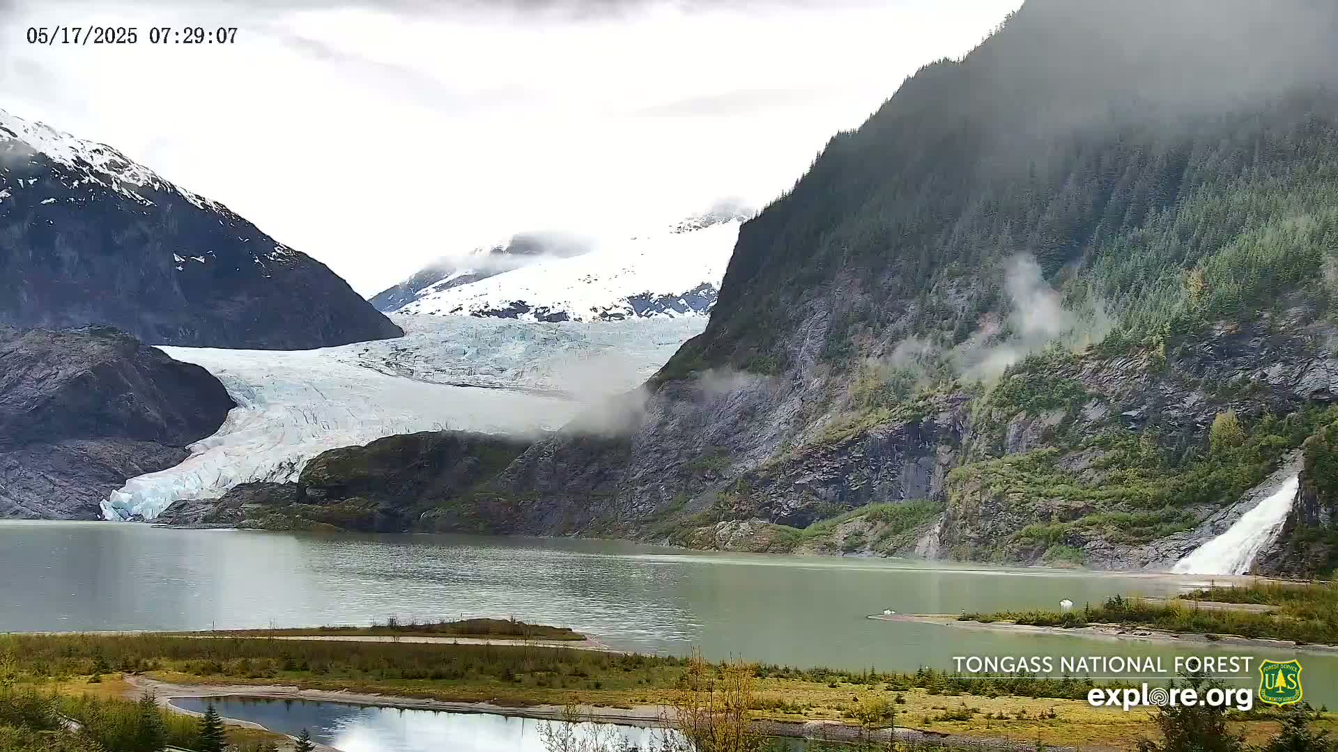 A glacier flows into a calm lake nestled between steep, mist-covered mountains, one of which features a waterfall.