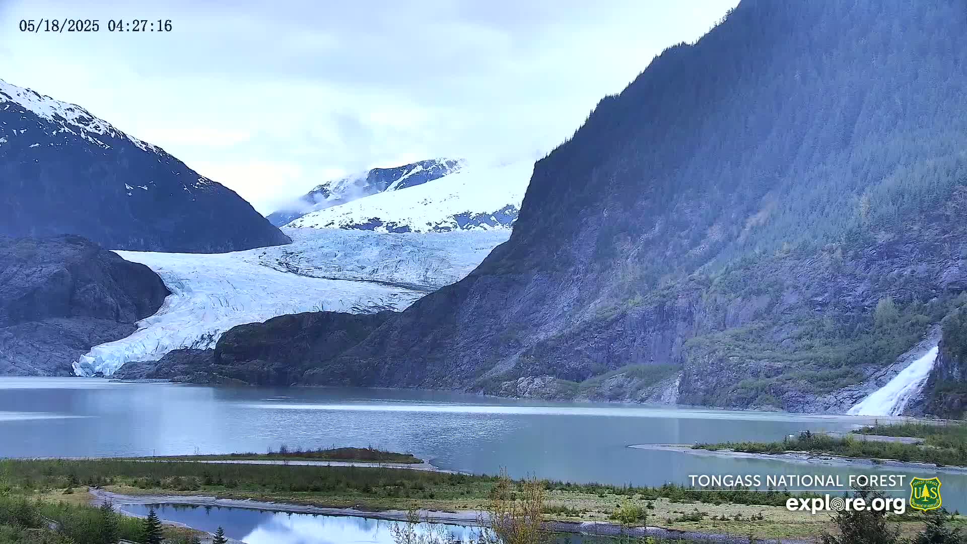 A large glacier flows into a calm lake, nestled between towering, dark mountains under a mostly cloudy sky.