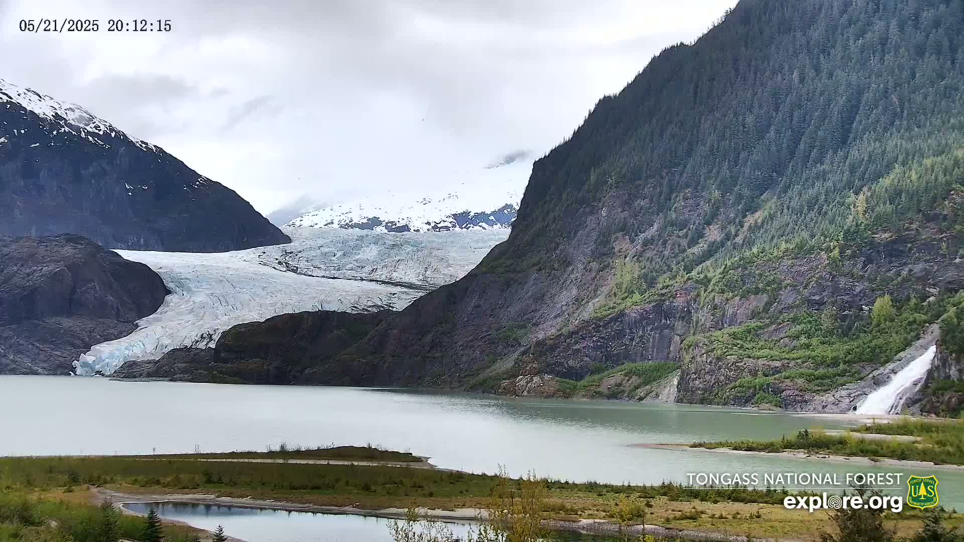 A glacier flows into a calm lake surrounded by mountains under an overcast sky.