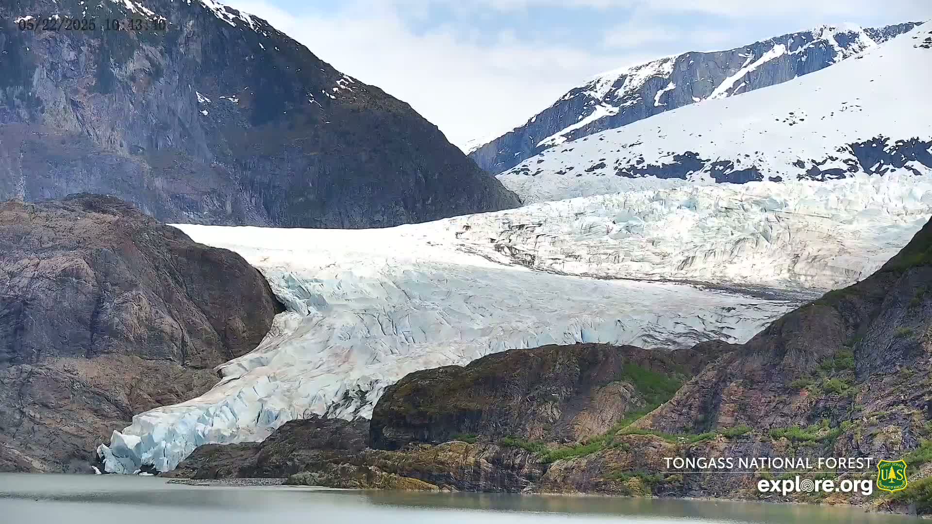 A large glacier flows down a mountainside into a calm body of water under a partly cloudy sky.