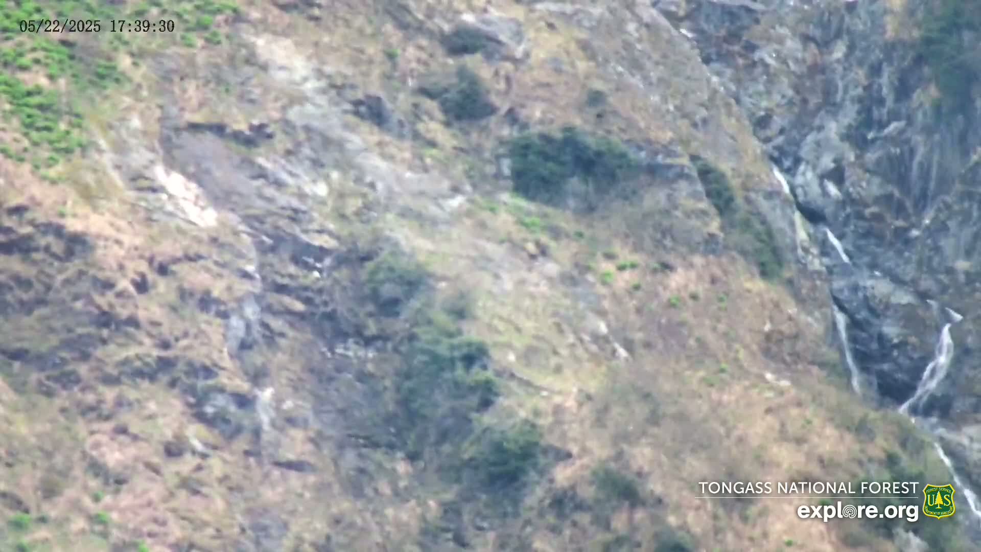 A rocky mountainside with sparse vegetation and several small waterfalls is visible, under what appears to be overcast conditions.