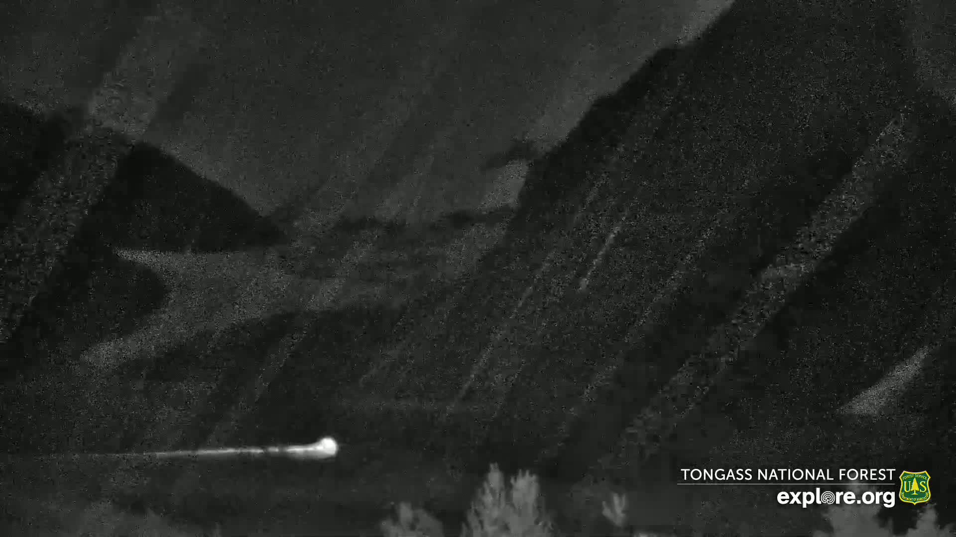A bright light streaks across a dark, mountainous landscape at night.