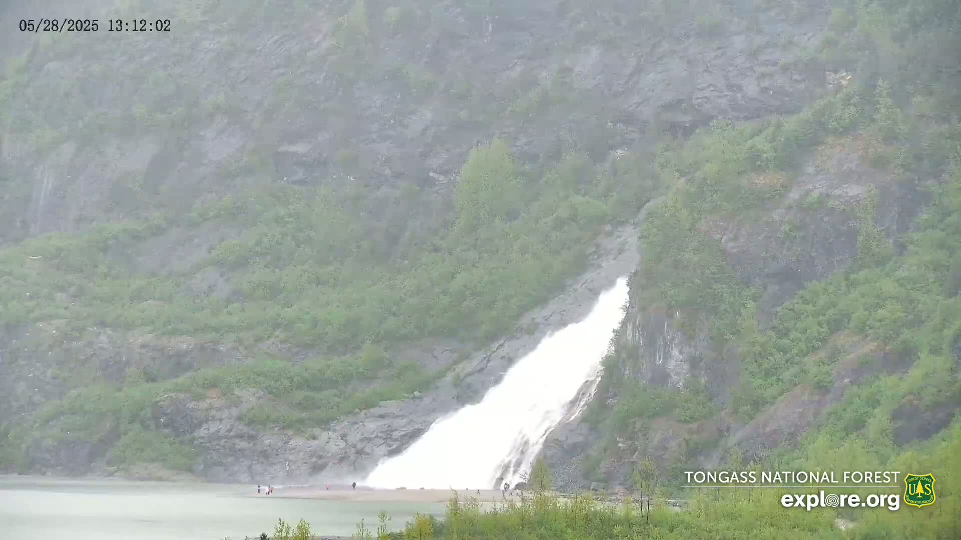 A large waterfall cascades down a rocky hillside into a lake under a heavy rain.