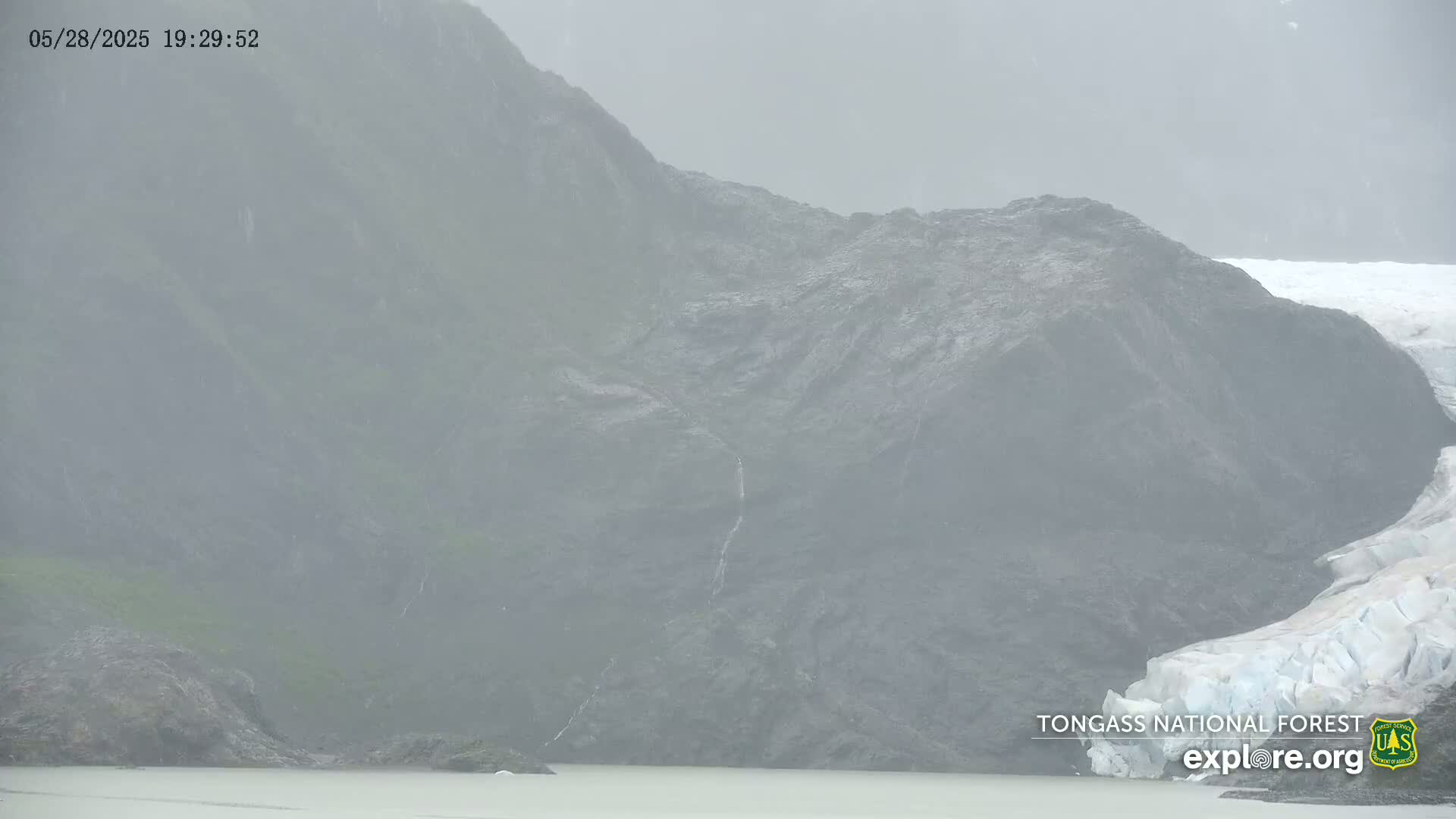 A rocky mountainside partially covered in fog and mist meets a glacier beside a body of still water.