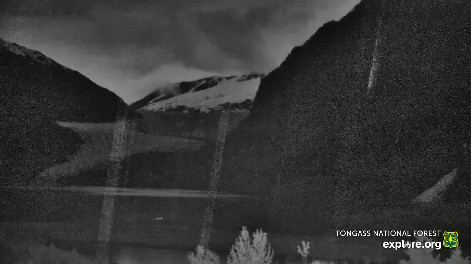 A dark, overcast night scene shows a calm body of water nestled between snow-dusted mountains.