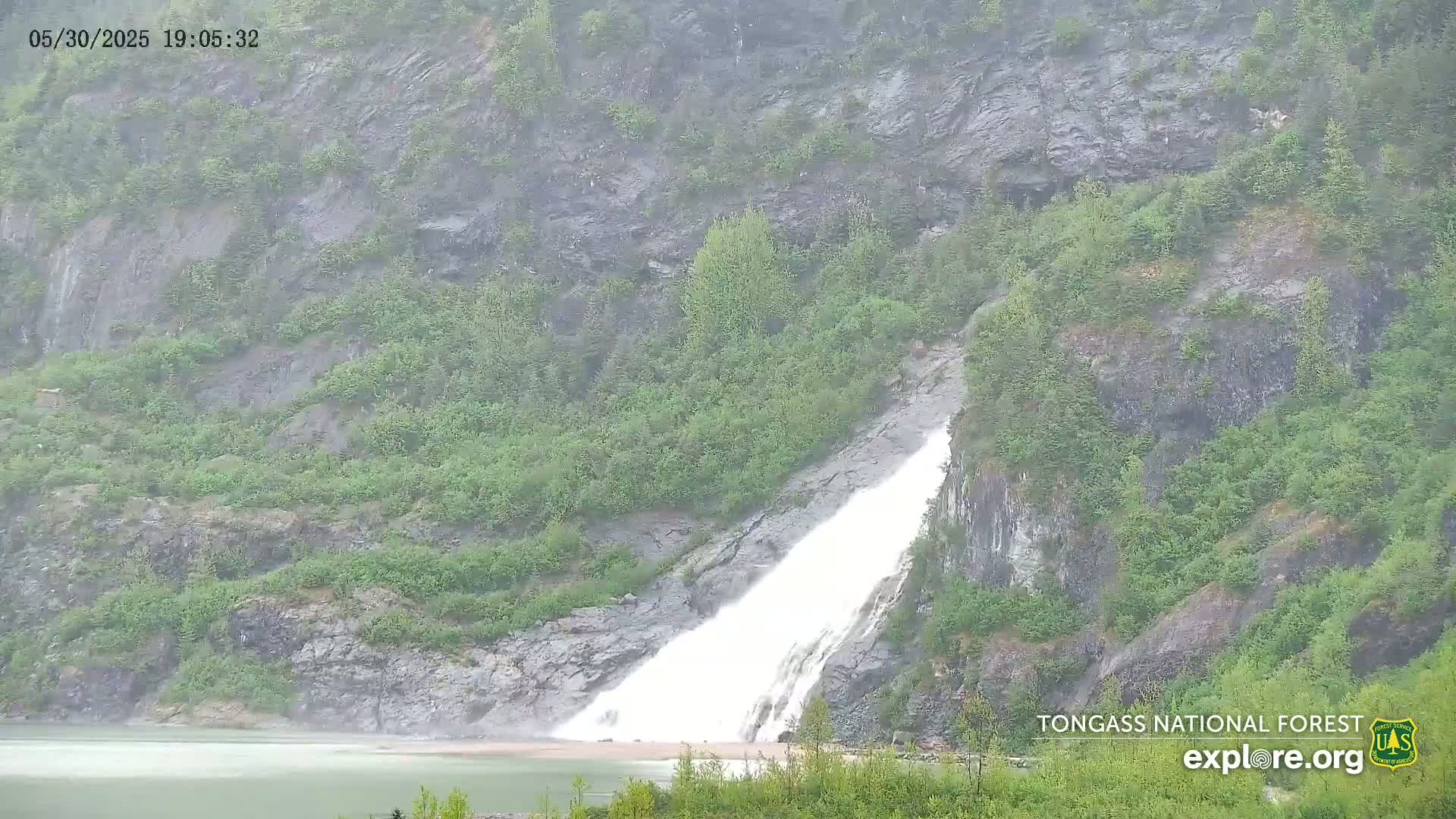 A large waterfall cascades down a rocky hillside covered in lush green vegetation during a rainy day.